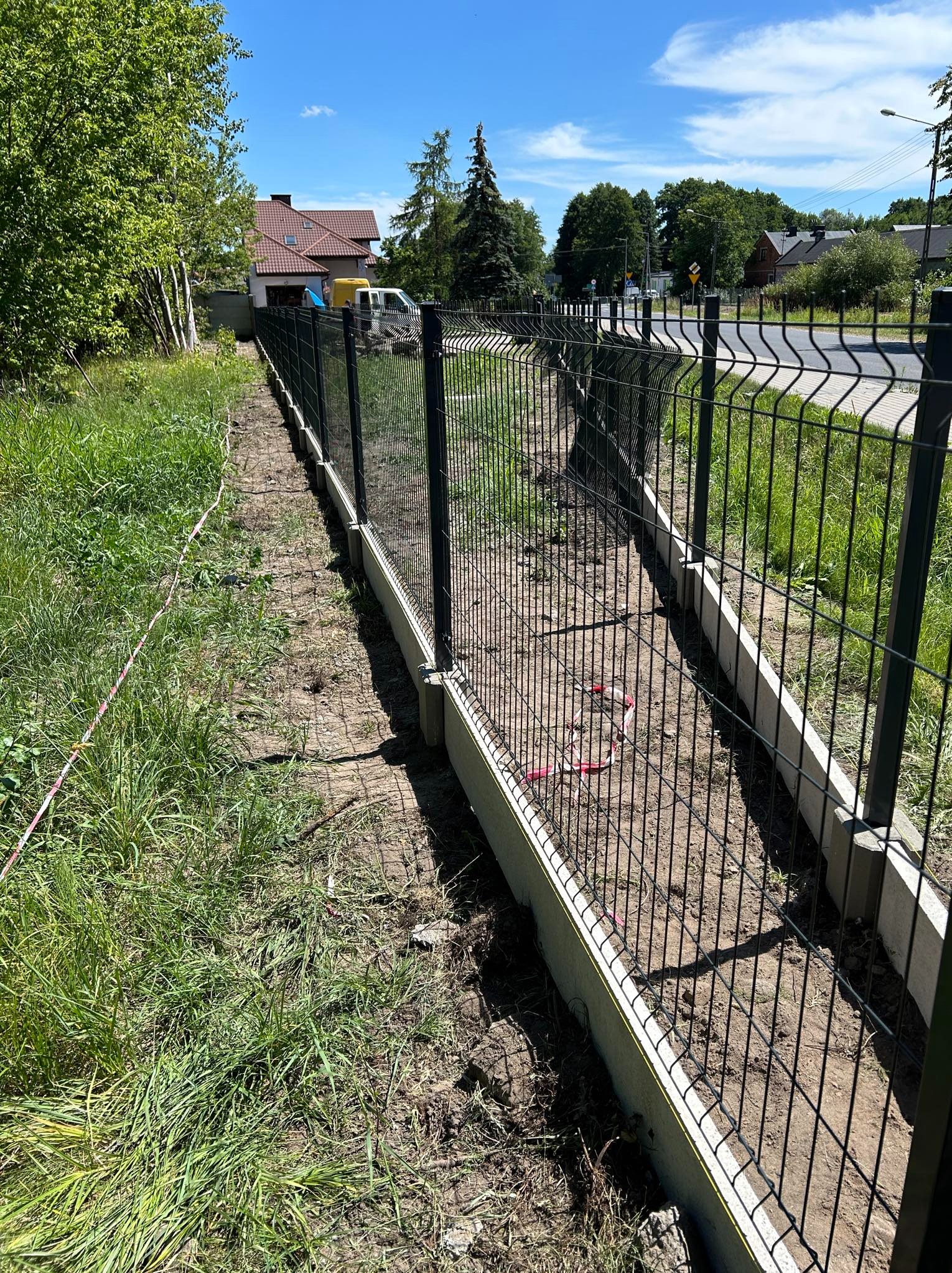 Nowo postawione, czarne ogrodzenie panelowe na betonowej podmurówce, widoczne z boku wzdłuż działki, z trawą po obu stronach i rozciągniętą czerwoną taśmą ostrzegawczą.