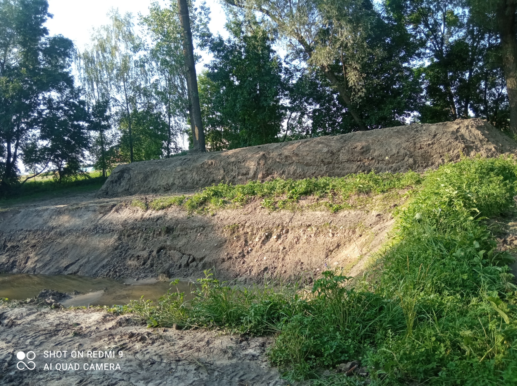 Dwa poziomy nasypów ziemnych w terenie z widocznymi śladami prac ziemnych i młodą roślinnością, w tle drzewa.