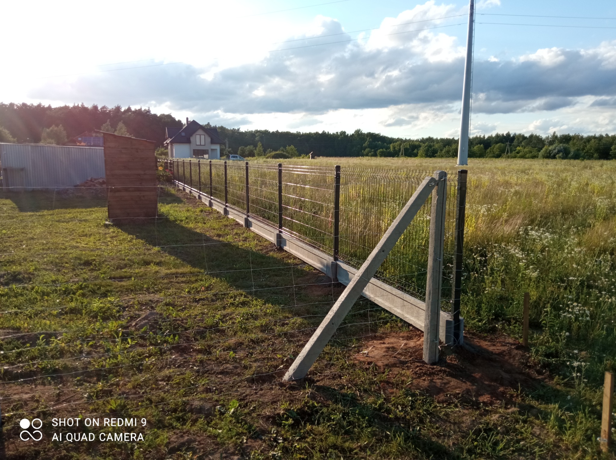 Montaż ogrodzenia panelowego na podmurówce betonowej z widocznym słupem podporowym, w tle łąka i dom.