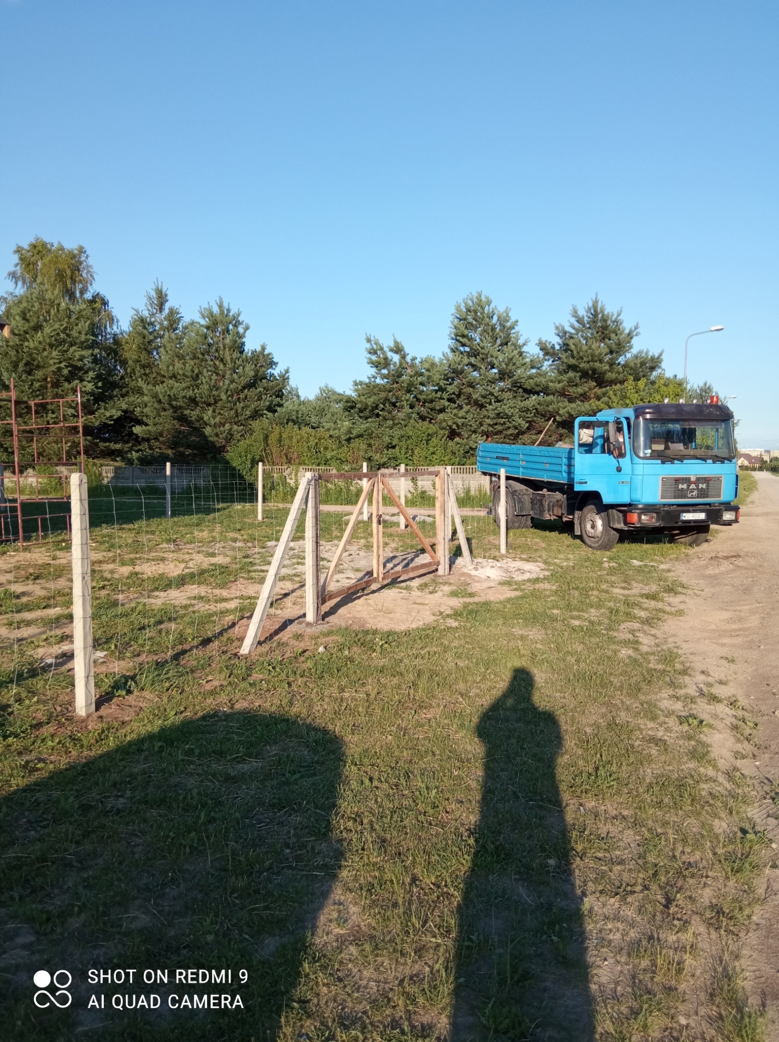 W trakcie montażu ogrodzenia z betonowych słupków i siatki, widoczny niebieski samochód ciężarowy MAN oraz cień osoby robiącej zdjęcie.
