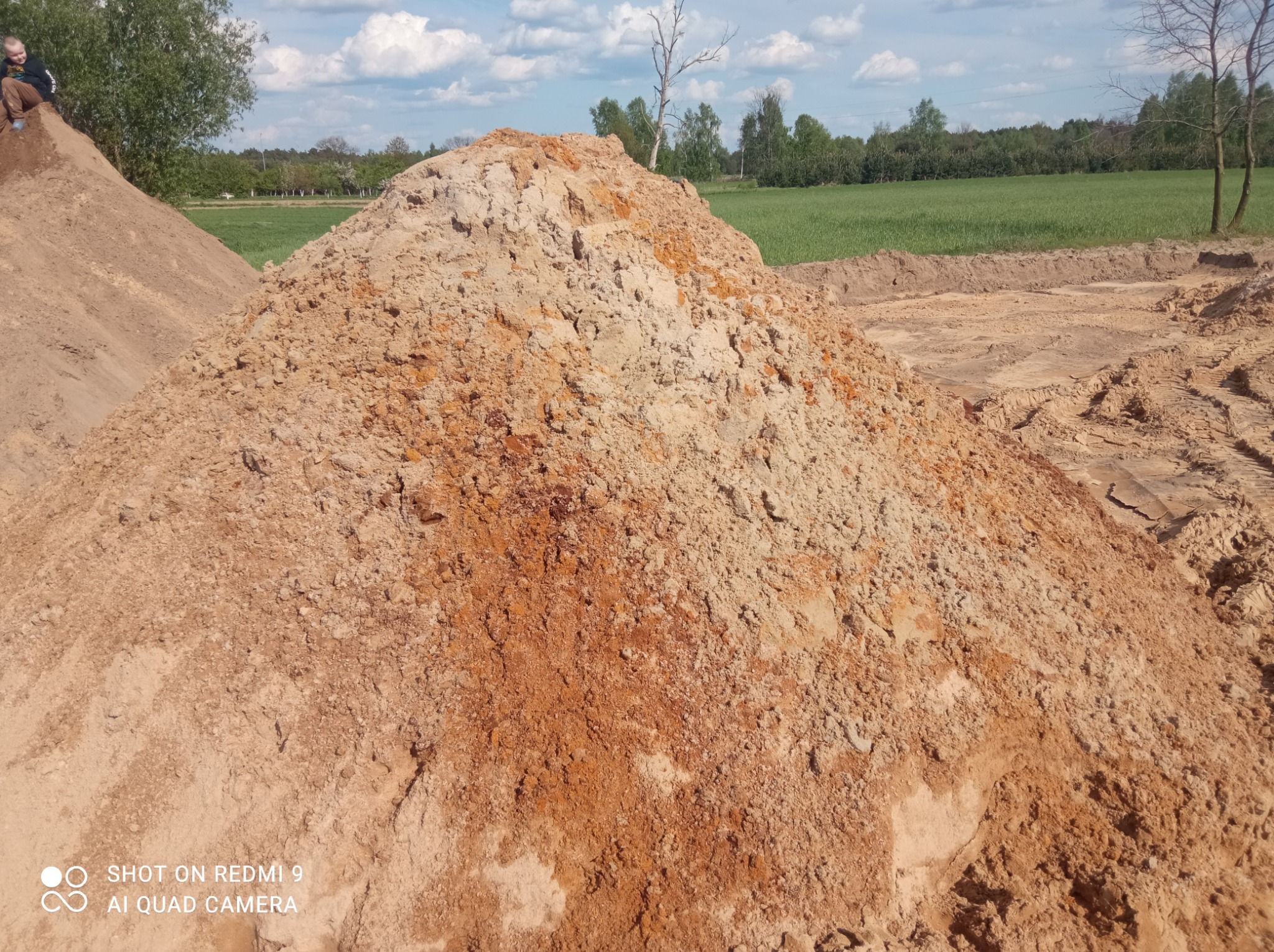Kupa piasku i ziemi na placu budowy, z widocznym wykopem w tle i osobą siedzącą na szczycie innej pryzmy ziemi.