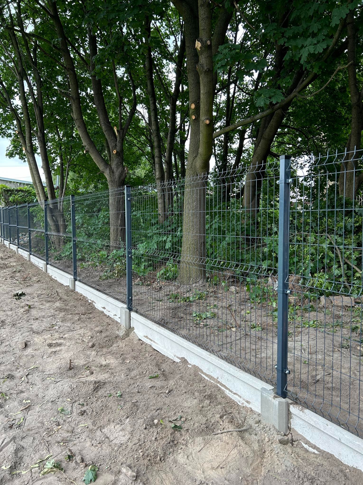 Nowo zainstalowane, ciemnoszare ogrodzenie panelowe z podmurówką betonową na tle drzew z przyciętymi gałęziami i zieleni, widok z poziomu gruntu na lekko nierównym terenie.