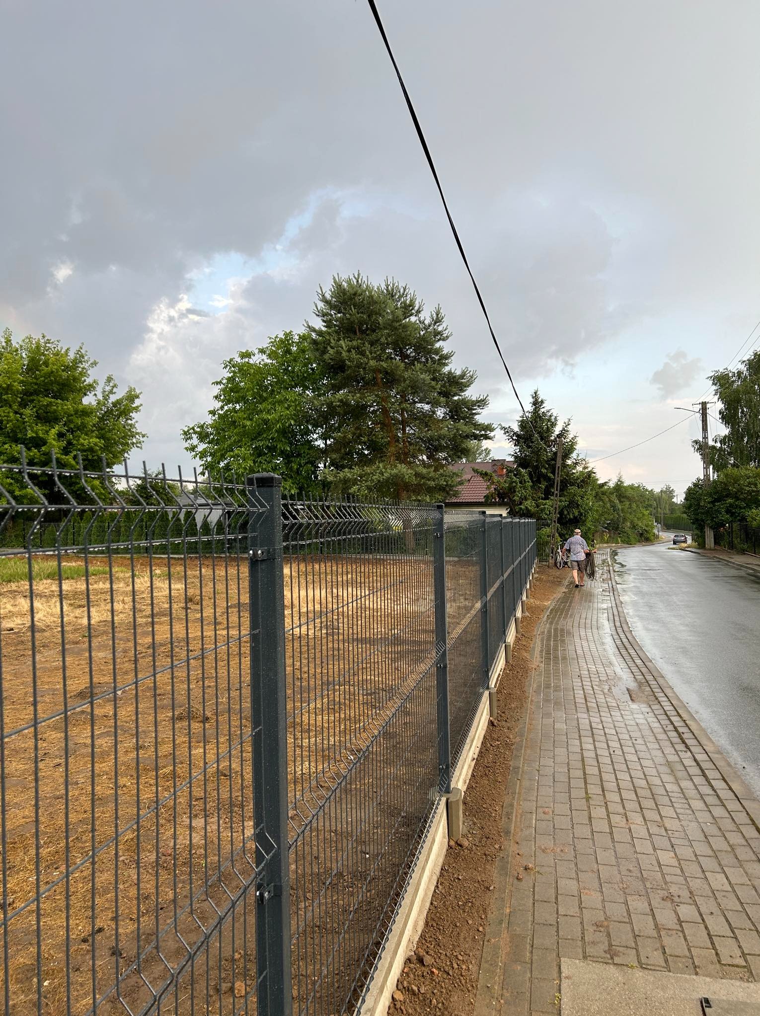 Nowo zamontowane, ciemnoszare ogrodzenie panelowe z betonową podmurówką, oddzielające działkę z suchą trawą od chodnika z kostki, z osobą idącą w oddali i pochmurnym niebem w tle.