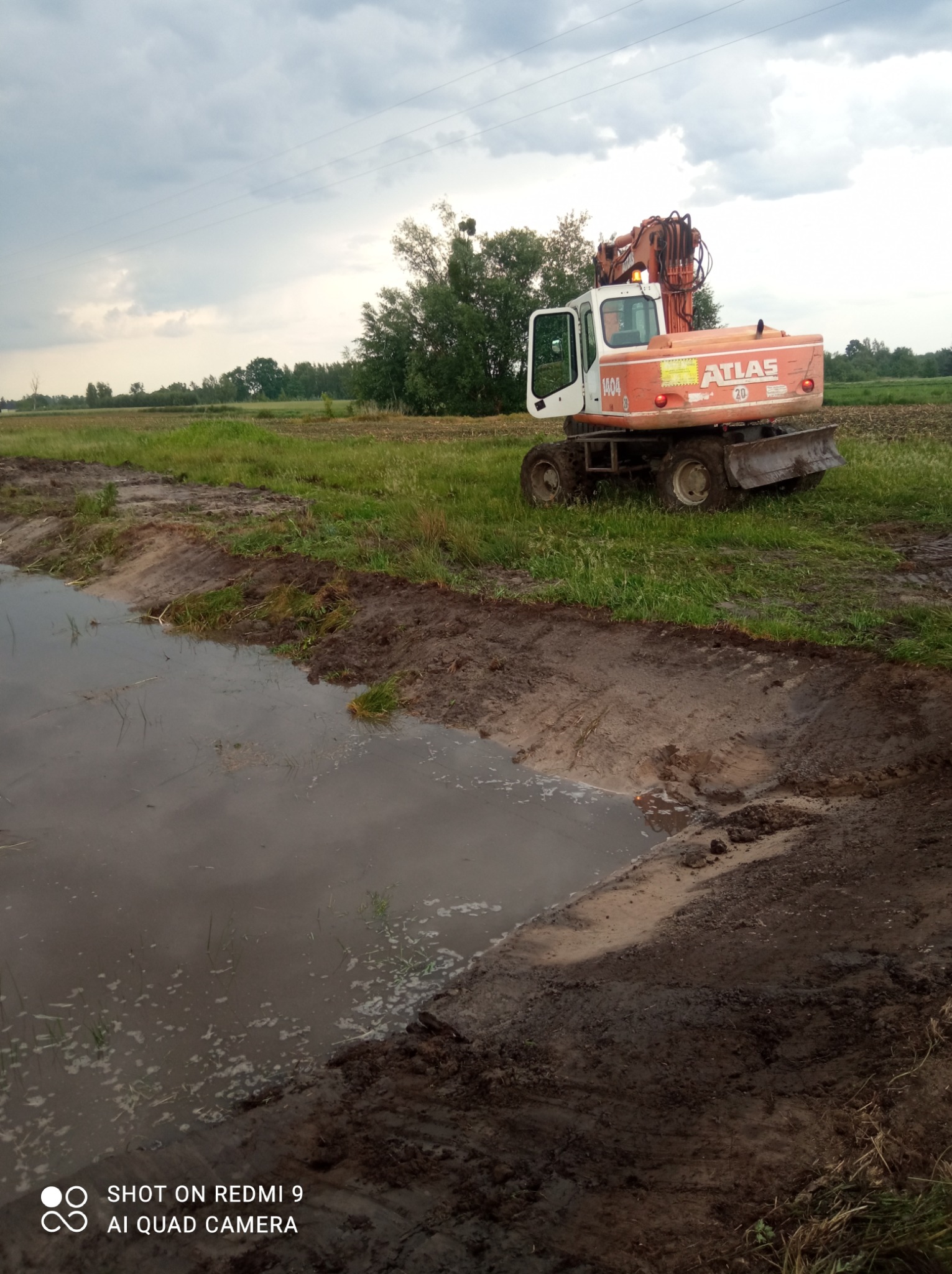 Pomarańczowo-biała koparka Atlas 1404 stoi na brzegu wykopu wypełnionego wodą na tle łąki i pochmurnego nieba.