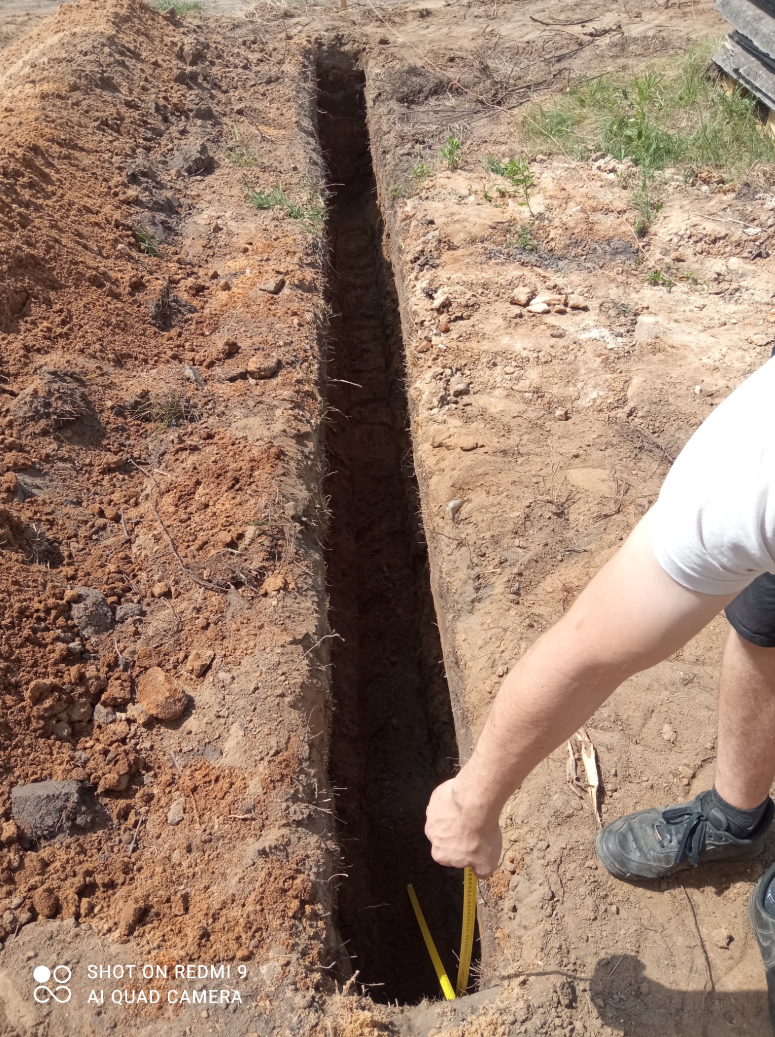 Wąski wykop w ziemi, mierzony żółtą taśmą mierniczą przez osobę w białej koszulce i czarnych butach, widok z góry, ziemia wokół wykopu nierówna.