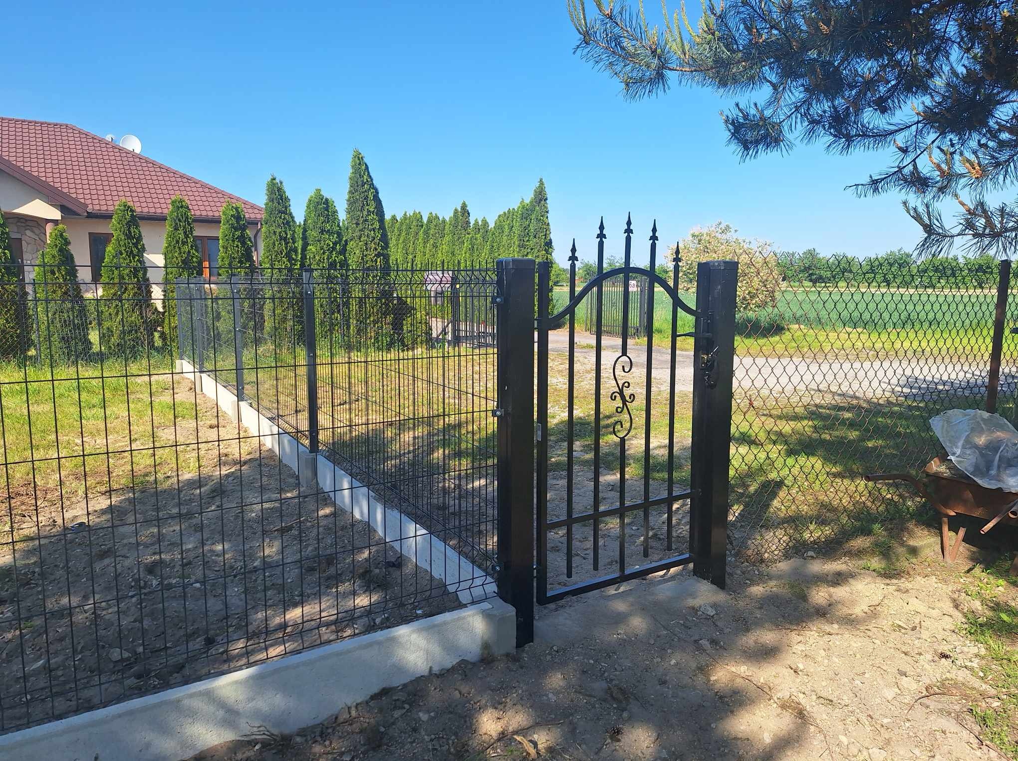 Czarna metalowa furtka z ozdobnymi elementami w ogrodzeniu panelowym na betonowej podmurówce, widok na dom i tuje w tle, wózek budowlany po prawej stronie.