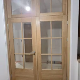 Dwu-skrzydłowe drzwi wewnętrzne z jasnego drewna z szybami, widoczne ślady po remoncie, w tle rura owinięta folią.