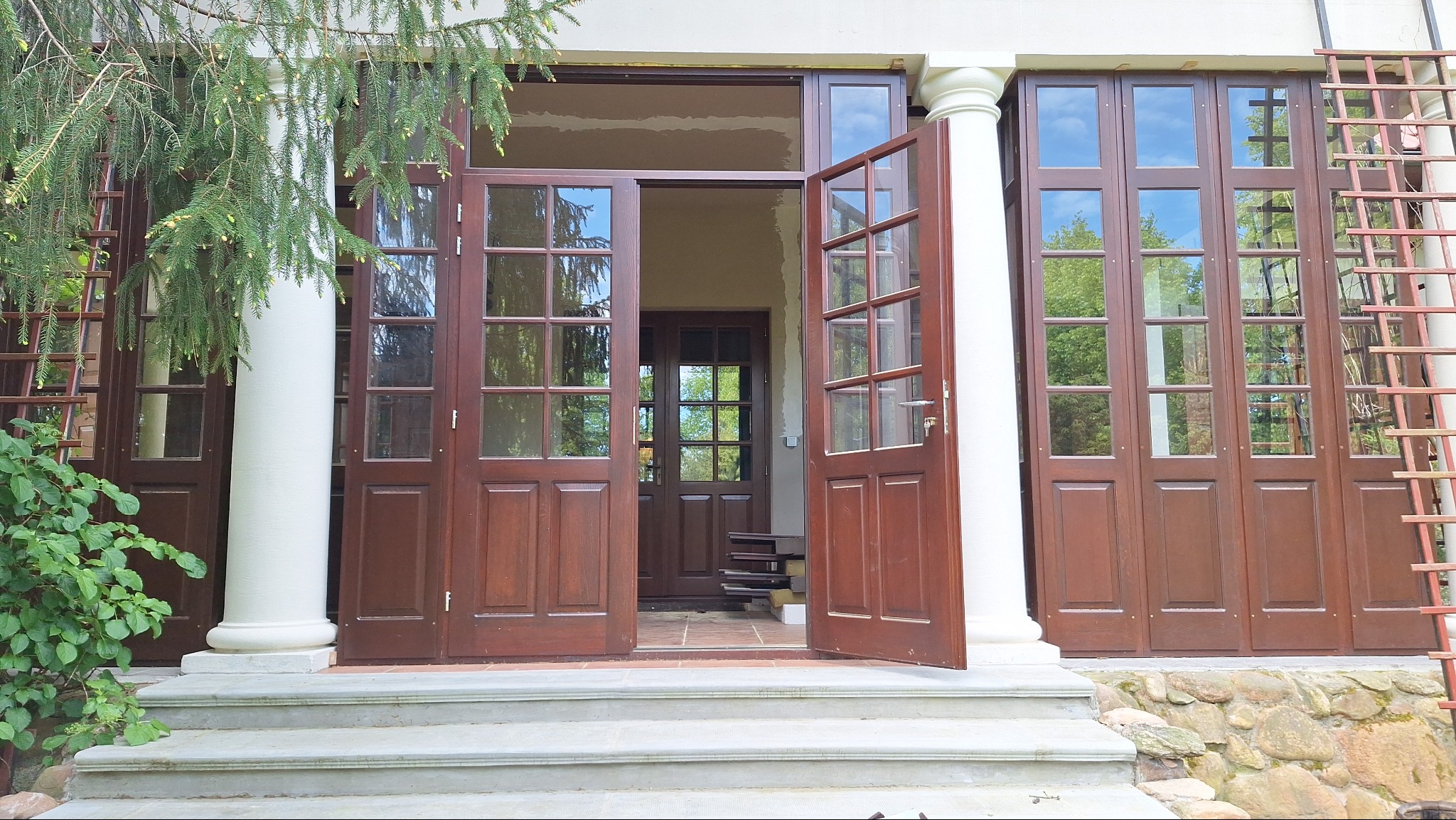 Wejście do domu w Wasilkowie z szerokimi, przeszklonymi drzwiami w kolorze ciemnego drewna, otwarte skrzydło ukazuje wnętrze, a białe kolumny dodają elegancji.