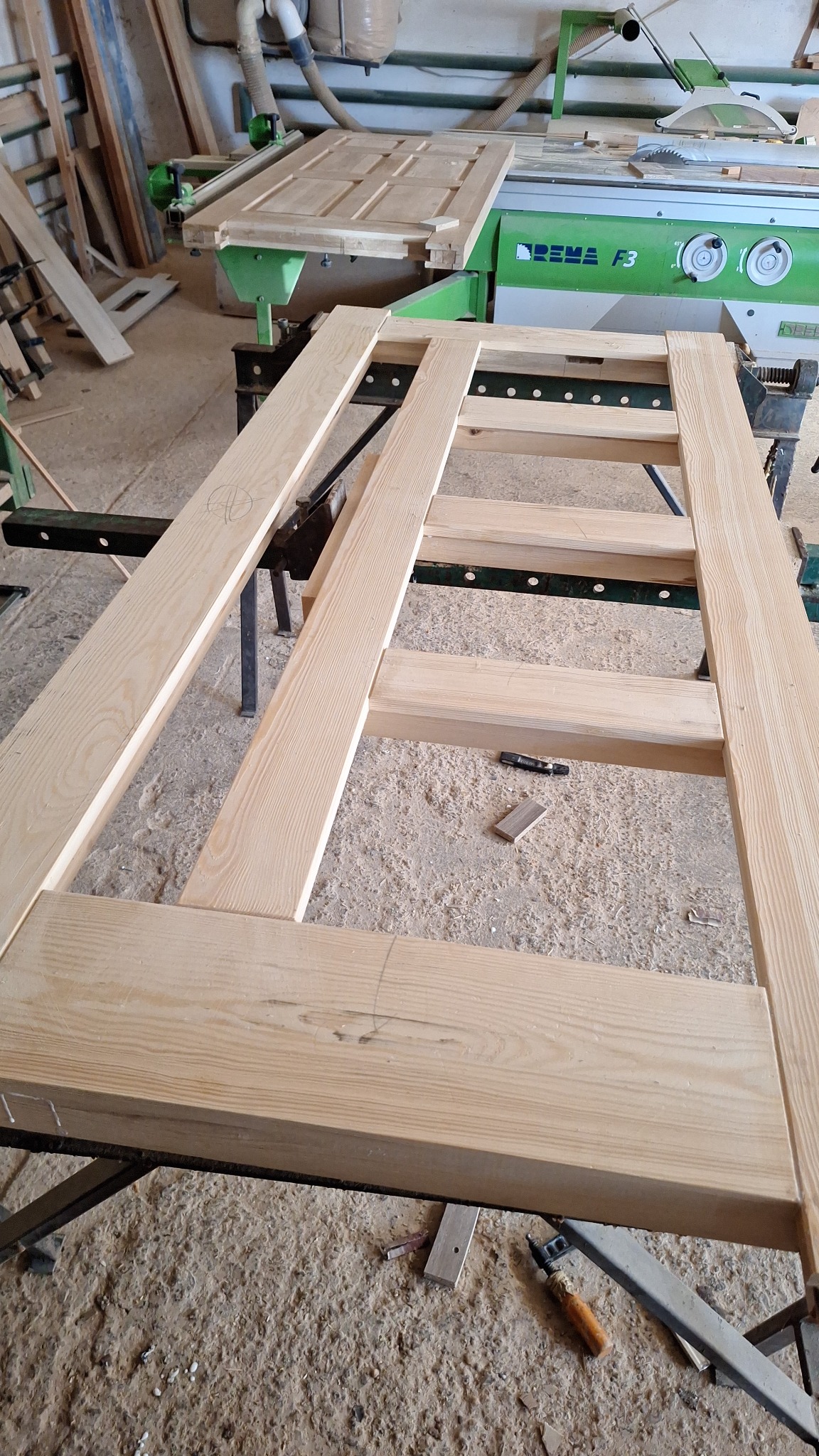 Rama drewnianych drzwi w warsztacie stolarskim, widoczna piła stołowa i narzędzia stolarskie.