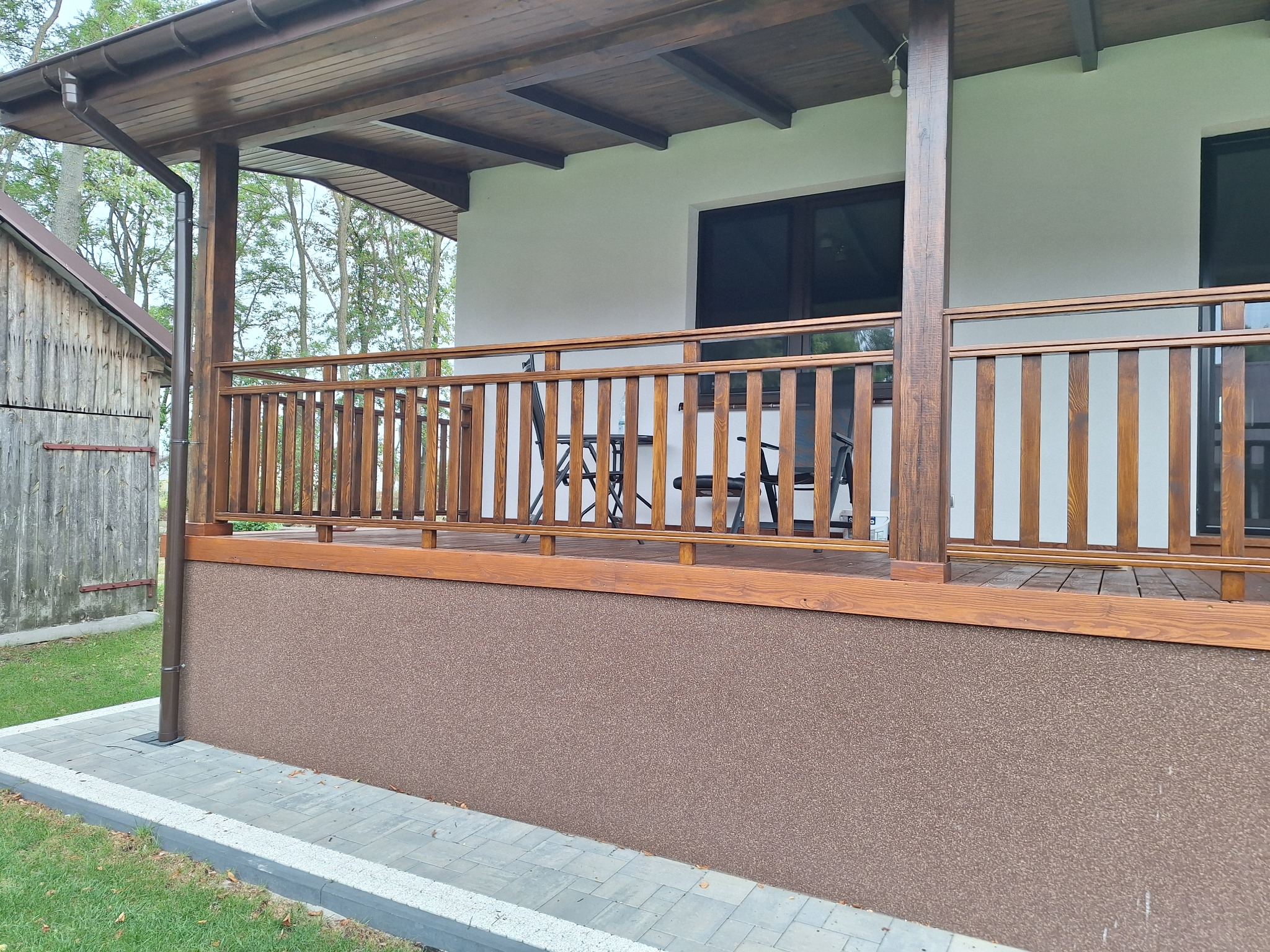 Zadaszony taras z drewnianą balustradą i podłogą, z widocznym stołem i krzesłami w tle, z brązową elewacją i widocznym budynkiem gospodarczym obok.