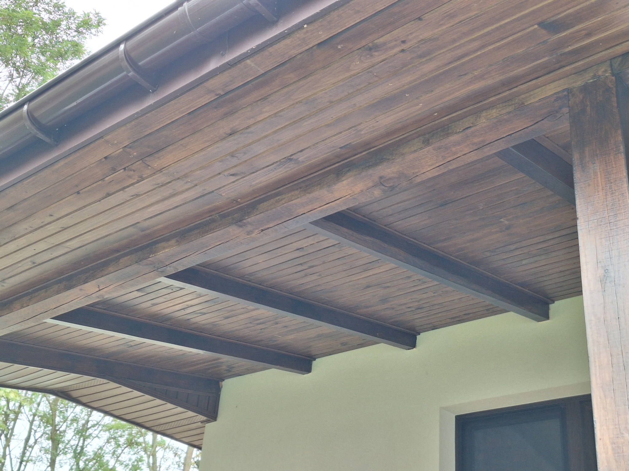 Zadaszenie tarasu z ciemnego drewna, widok od dołu, widoczne krokwie i rynna, ściana budynku w tle.