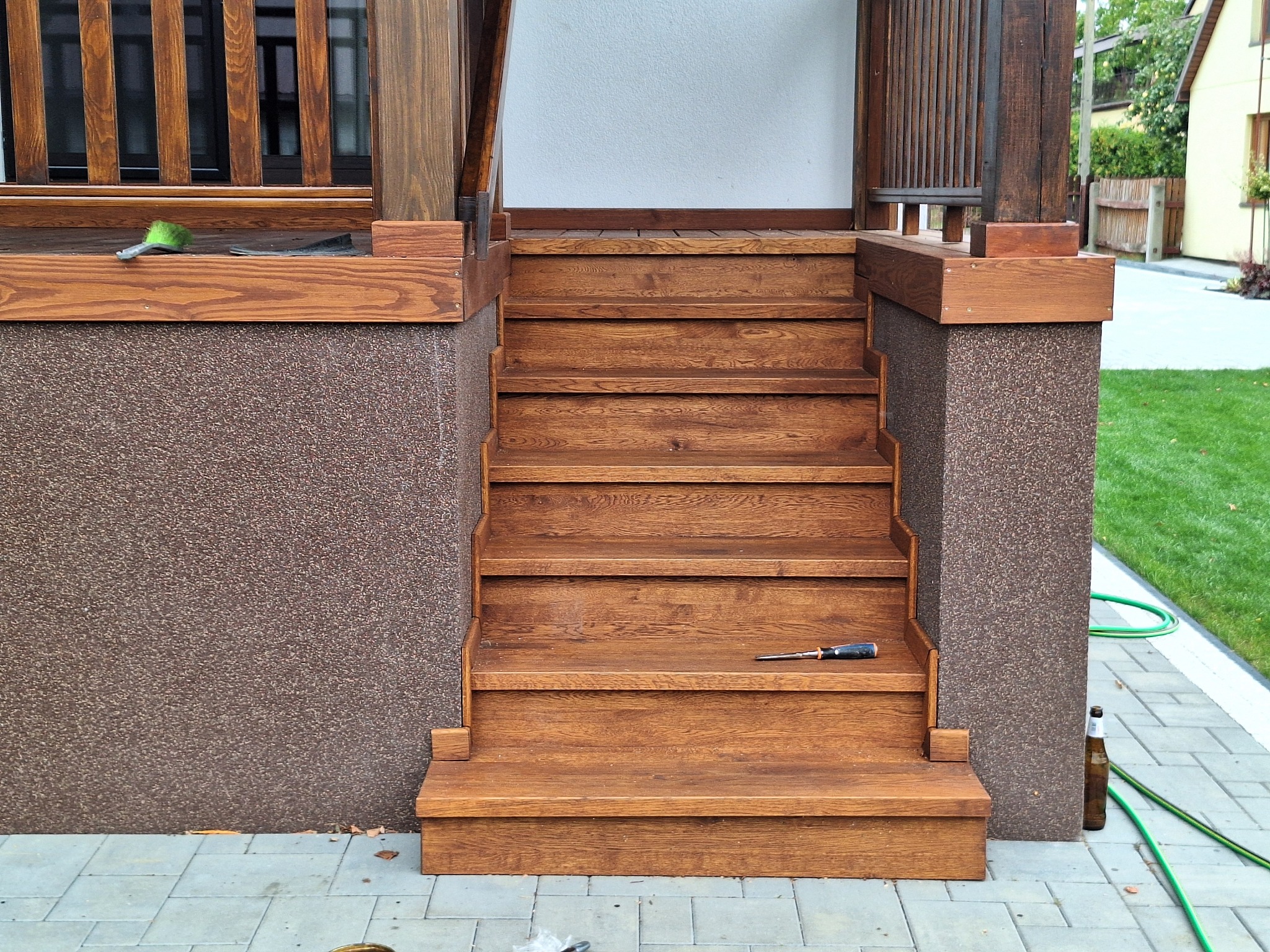 Zewnętrzne, drewniane schody wejściowe z narzędziami i butelką po piwie, prowadzące do domu z elewacją w kolorze brązowym.