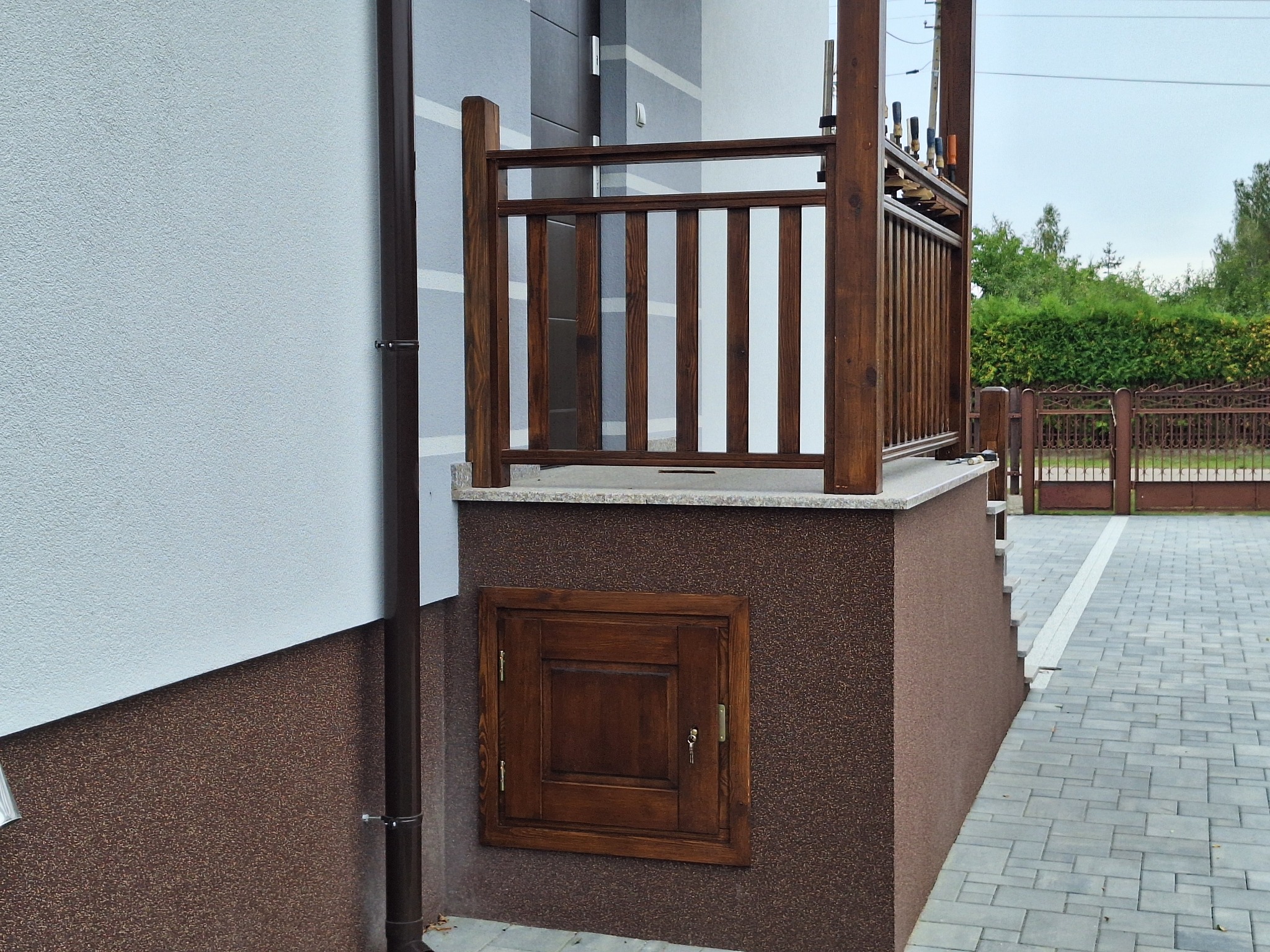 Fragment elewacji budynku z drewnianą balustradą werandy, małą klapą rewizyjną zamykaną na klucz i widocznymi śladami prac montażowych na balustradzie.