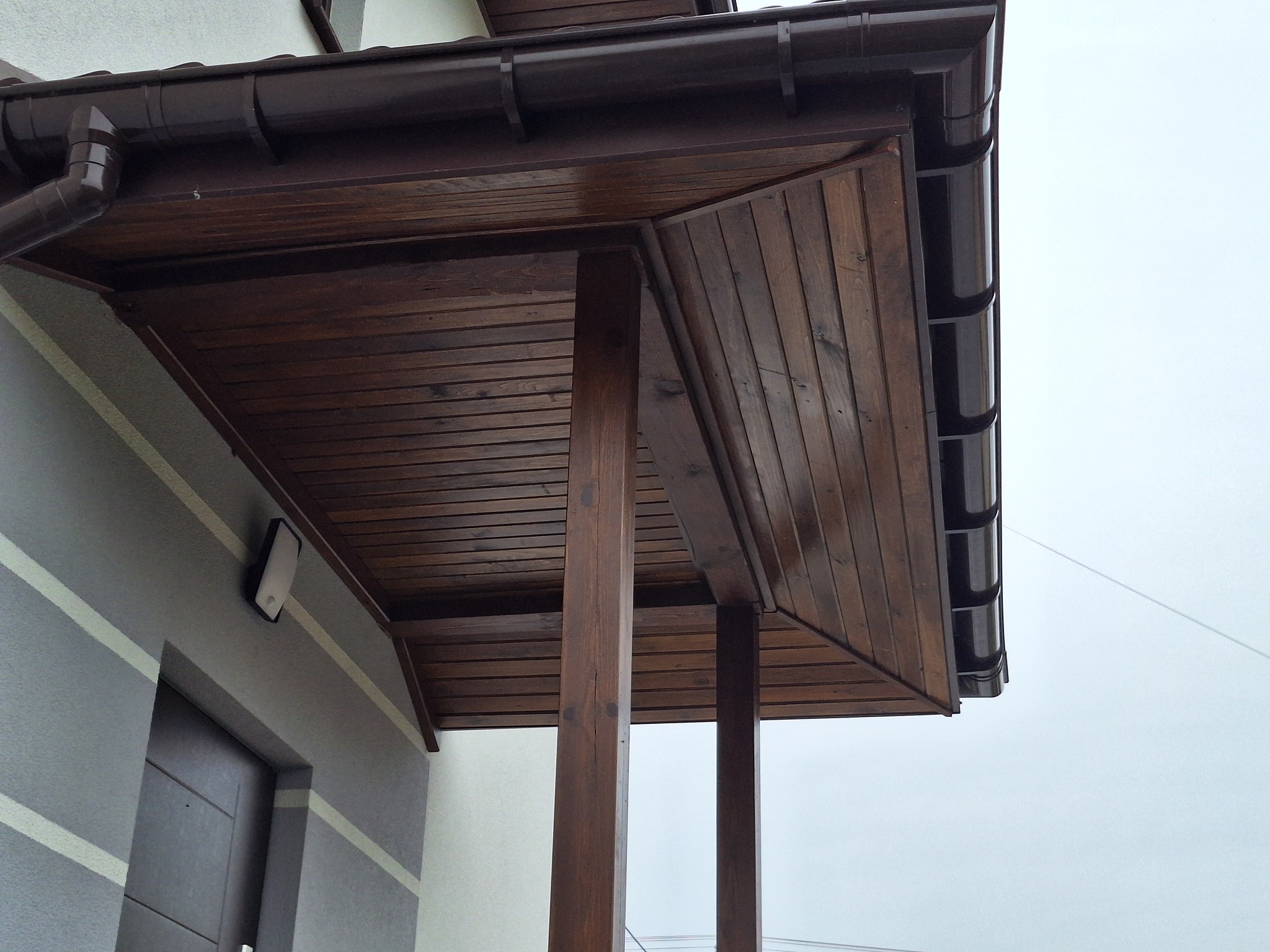 Zadaszenie wejścia do domu z brązowego drewna, wsparte na drewnianych słupach, z widoczną rynną i fragmentem elewacji z poziomymi pasami.