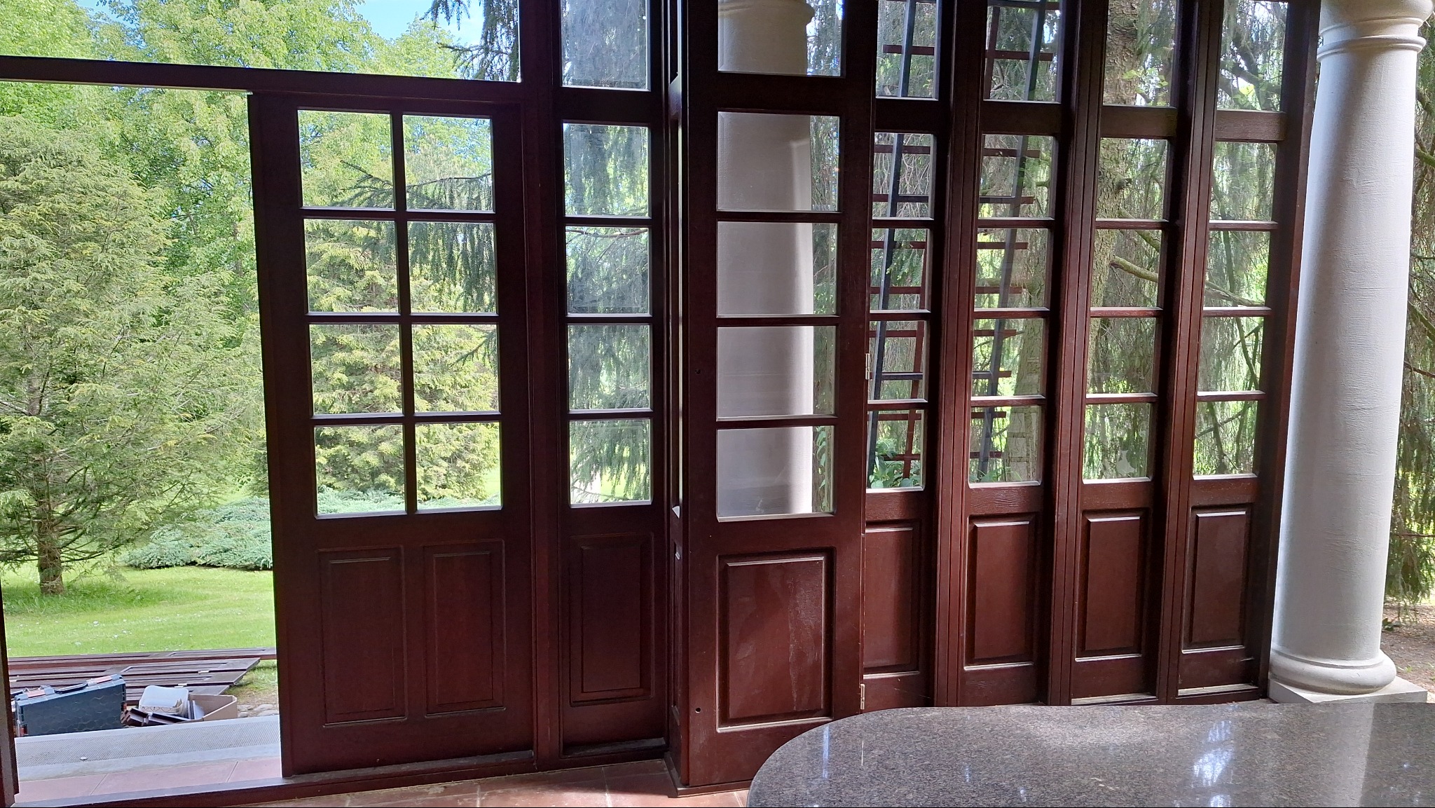 Rozsuwane, przeszklone drzwi w kolorze ciemnego brązu z widokiem na zielony ogród i białą kolumnę werandy.