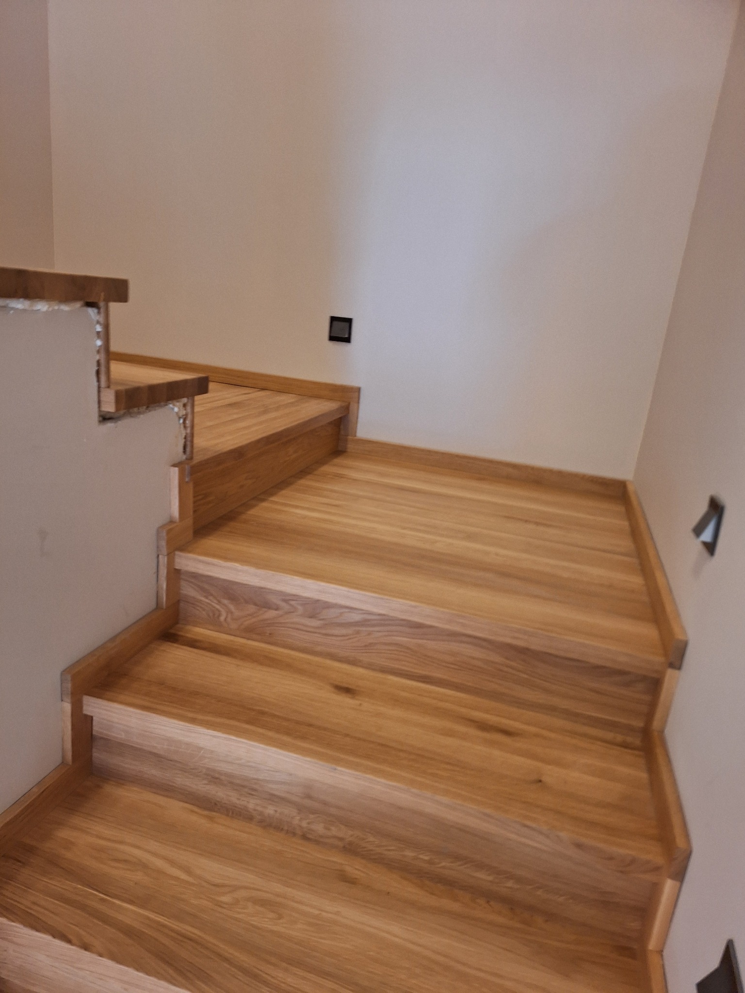 Surowe drewniane schody w trakcie montażu, widoczne niedokończone połączenia ze ścianą i brak wykończenia, naturalne oświetlenie podkreśla strukturę drewna.