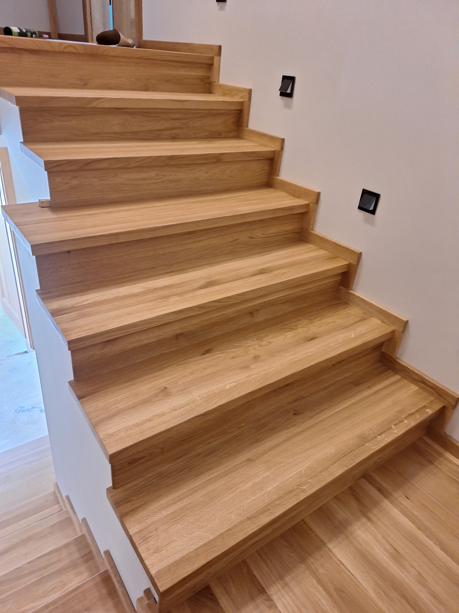 Jasne, dębowe stopnie schodów z minimalistycznymi, kwadratowymi lampkami ściennymi, widok z dołu.
