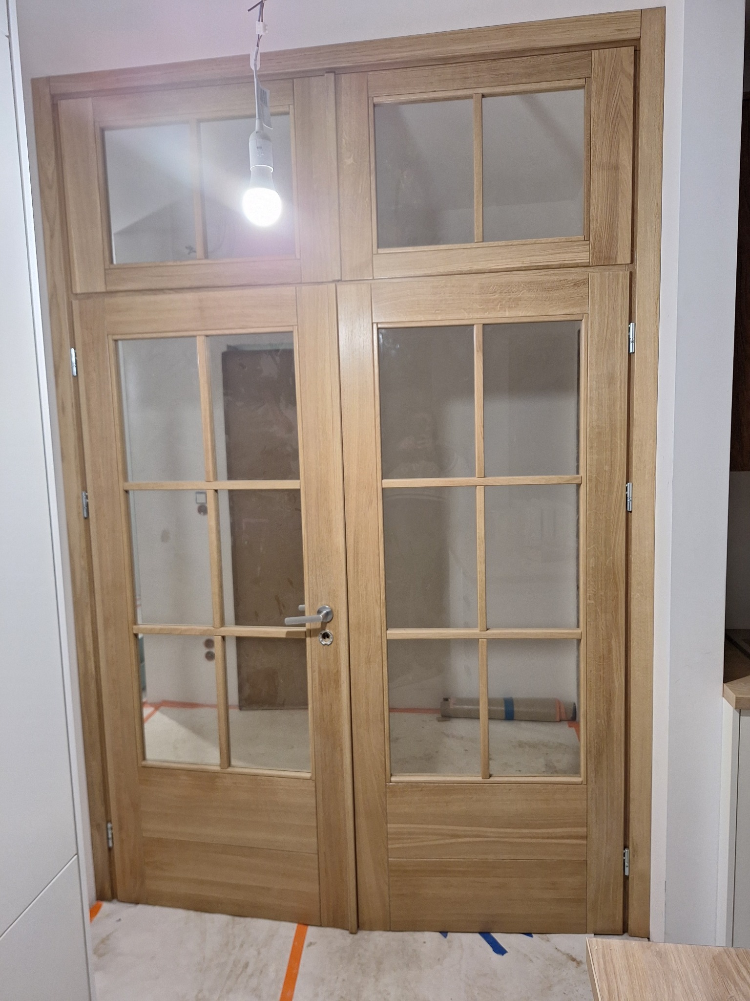 Dwu-skrzydłowe drzwi wewnętrzne z jasnego drewna z szybami, widoczne ślady po remoncie, w tle rura owinięta folią.