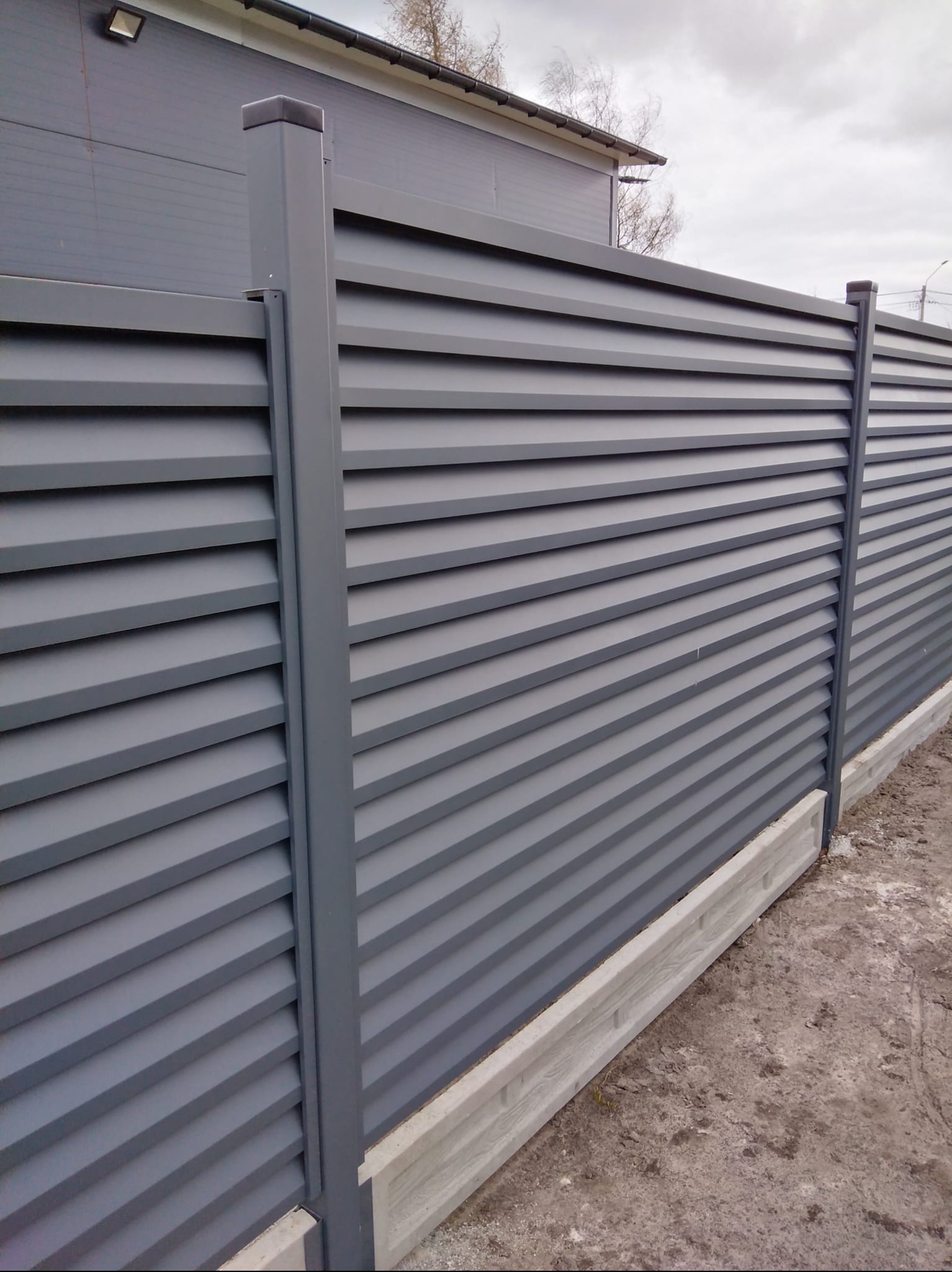 Nowoczesne ogrodzenie panelowe w odcieniach szarości z betonową podmurówką. Widoczne poziome lamele i pionowe słupki. Minimalistyczny design, stonowana kolorystyka.