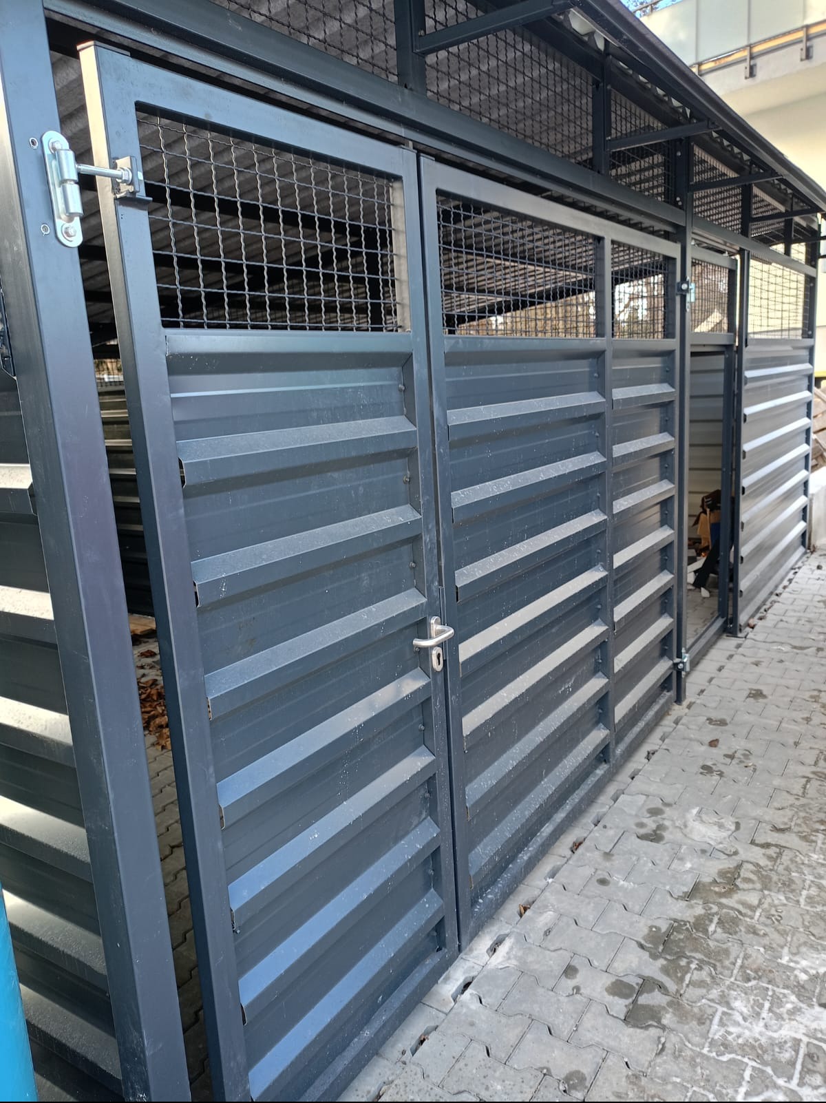 Metalowa konstrukcja z zamykanymi segmentami, prawdopodobnie na śmietniki, z widoczną kratą u góry i poziomymi panelami na drzwiach, na tle kostki brukowej.