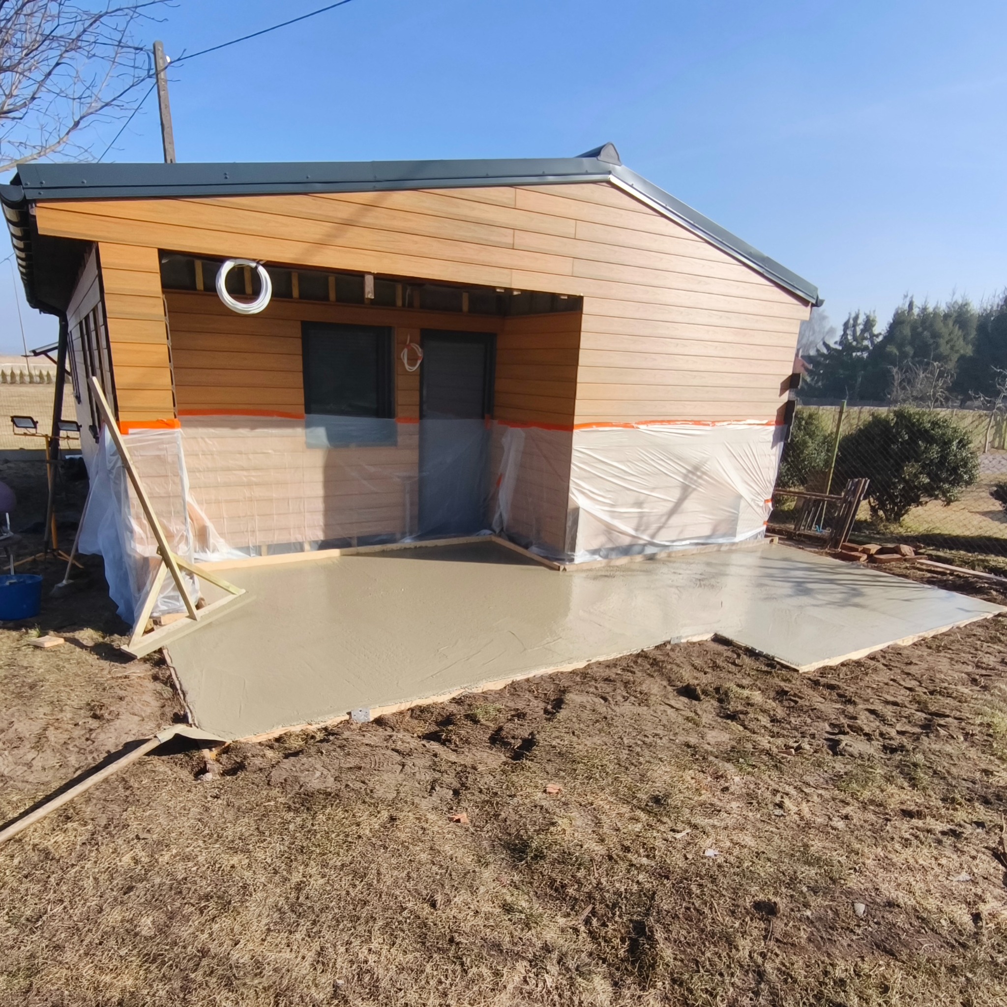 Wylewka betonowa przed domkiem z drewnianą elewacją, zabezpieczonym folią. Widoczne narzędzia i drewniane szalunki. Słoneczny dzień.