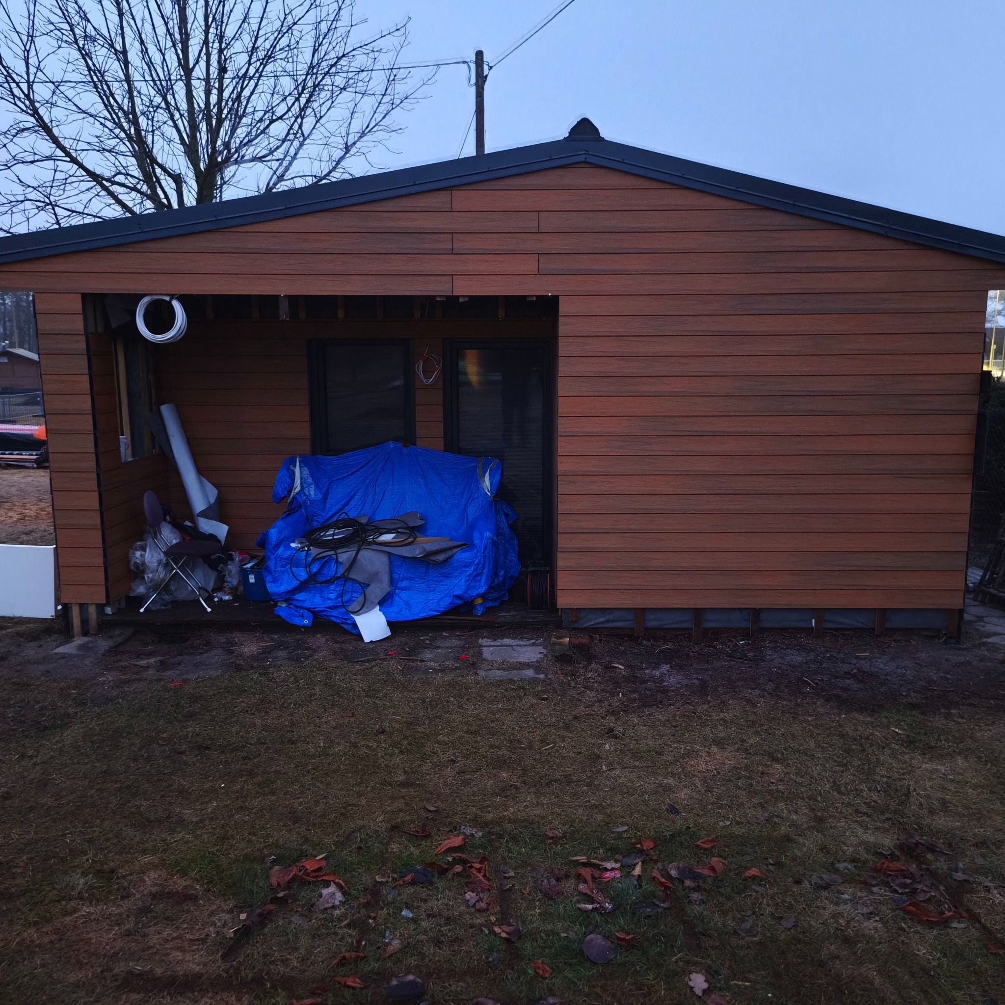 Niedokończona altana z brązową elewacją, przykryta niebieską plandeką. Widoczne narzędzia i materiały budowlane. Surowy krajobraz, jesienne liście na ziemi.
