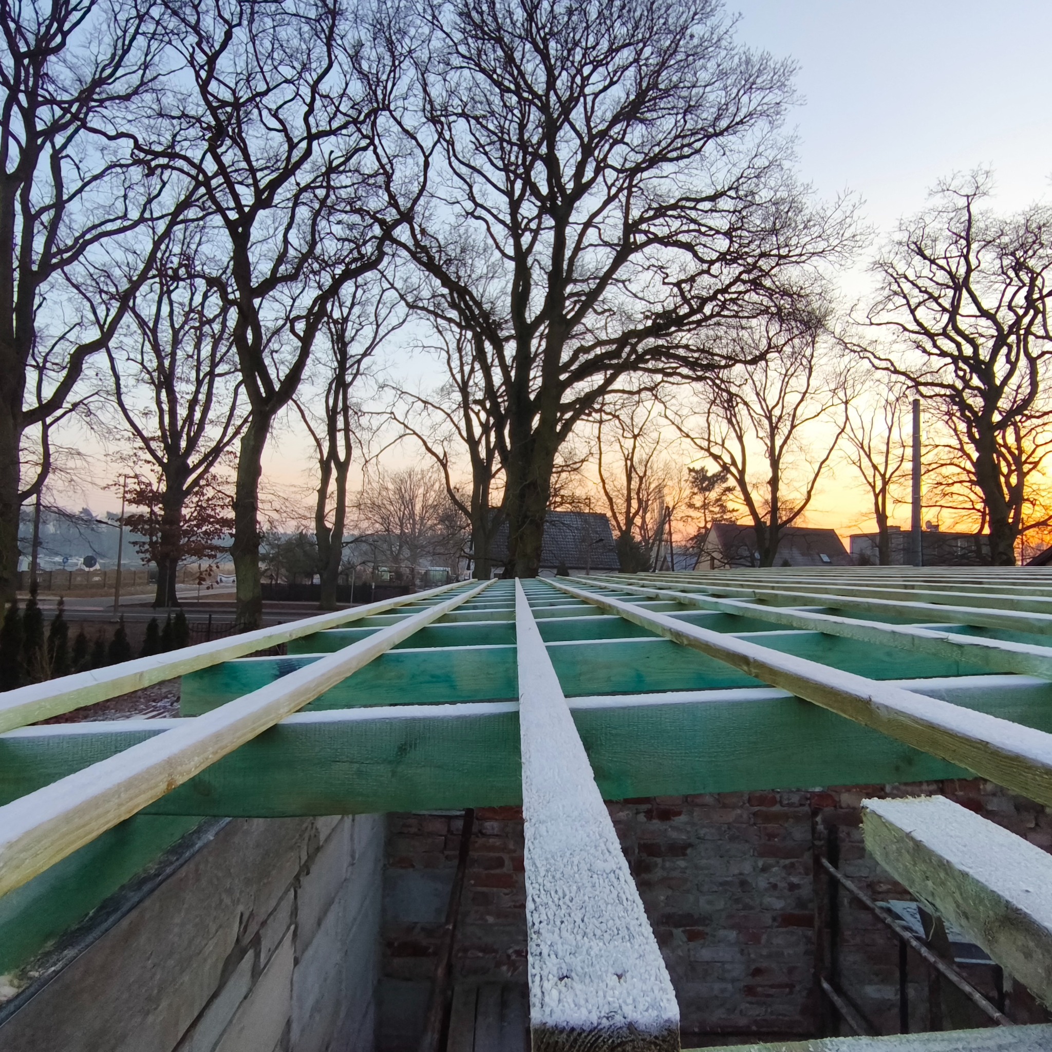 Konstrukcja dachu z drewnianych belek, pokrytych szronem, na tle zachodzącego słońca i drzew w Świnoujściu. Widok z wnętrza budynku.