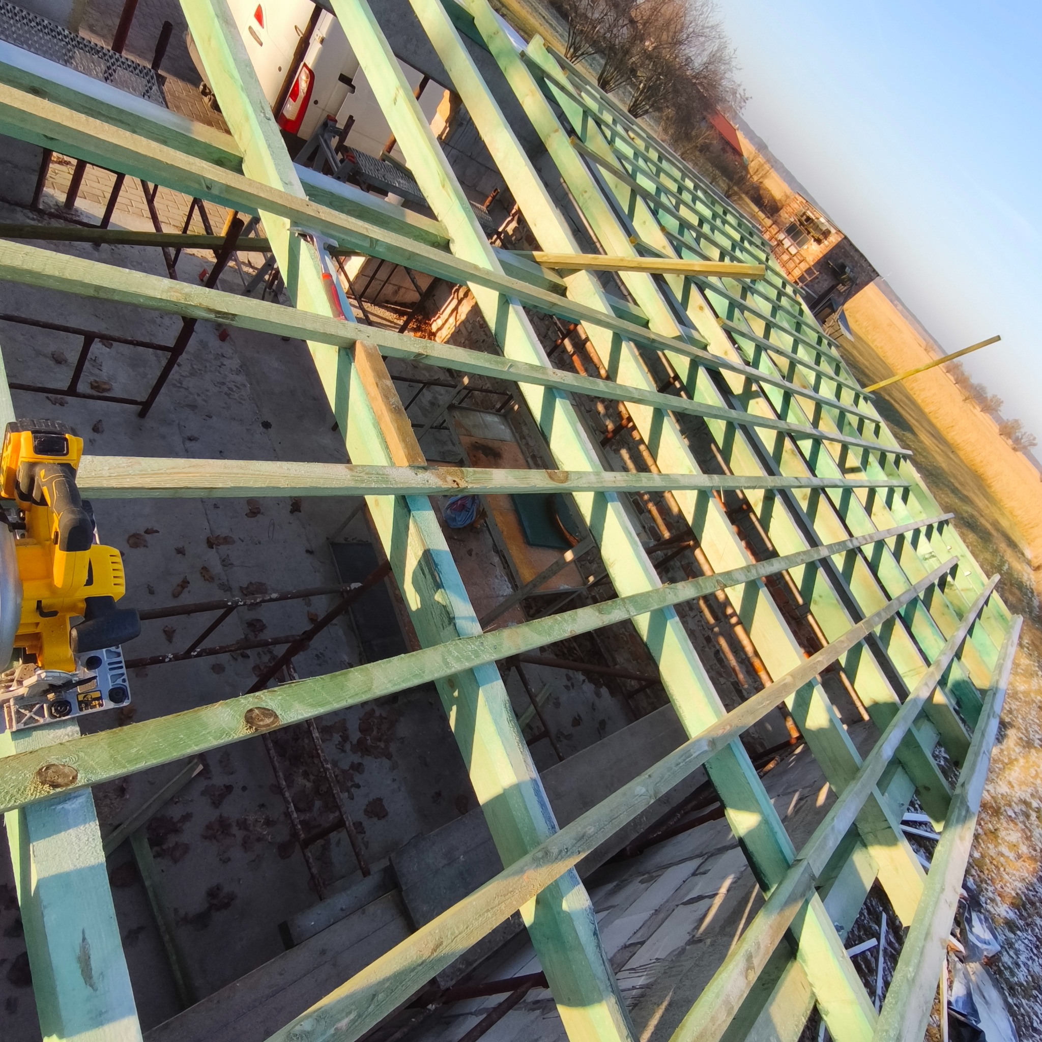 Konstrukcja dachu z impregnowanego drewna, widok z góry, z żółtą piłą tarczową po lewej. W tle budynek w trakcie budowy i pole. Ujęcie pod kątem.