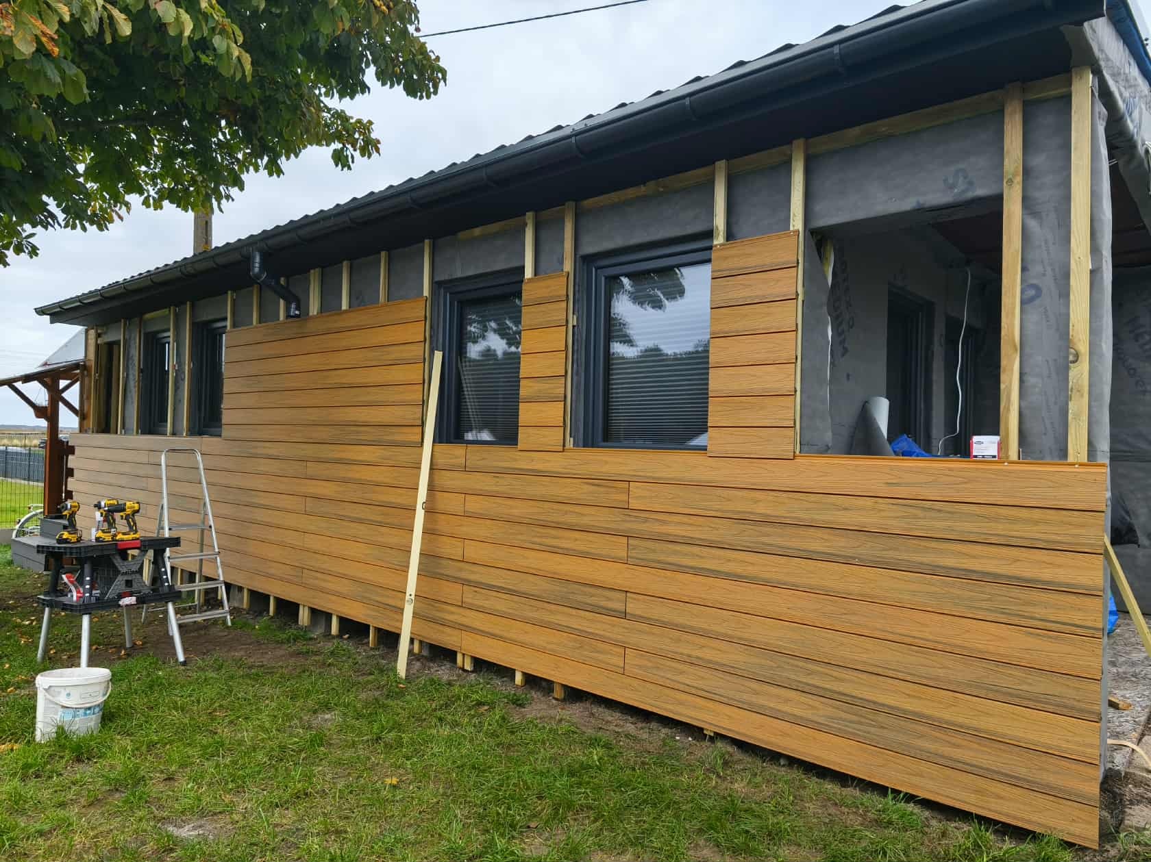 Montaż elewacji drewnopodobnej na budynku jednorodzinnym. Widoczne narzędzia pracy, fragment elewacji w trakcie montażu, okna. Ujęcie z zewnątrz, w dzień.