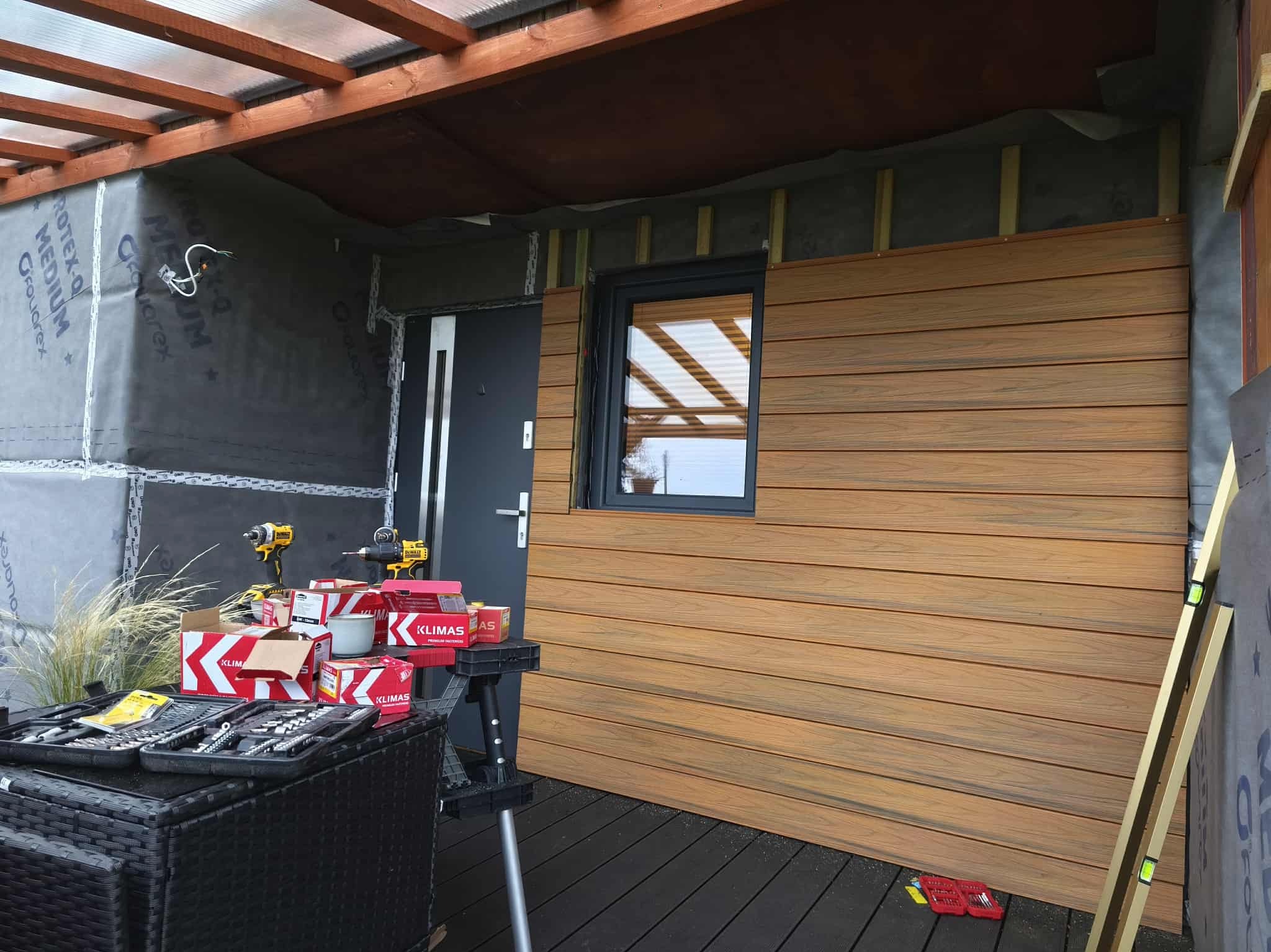 Montaż paneli elewacyjnych imitujących drewno na zewnątrz budynku. Widoczne narzędzia, poziomica i materiały montażowe. Częściowo wykończona ściana z oknem.