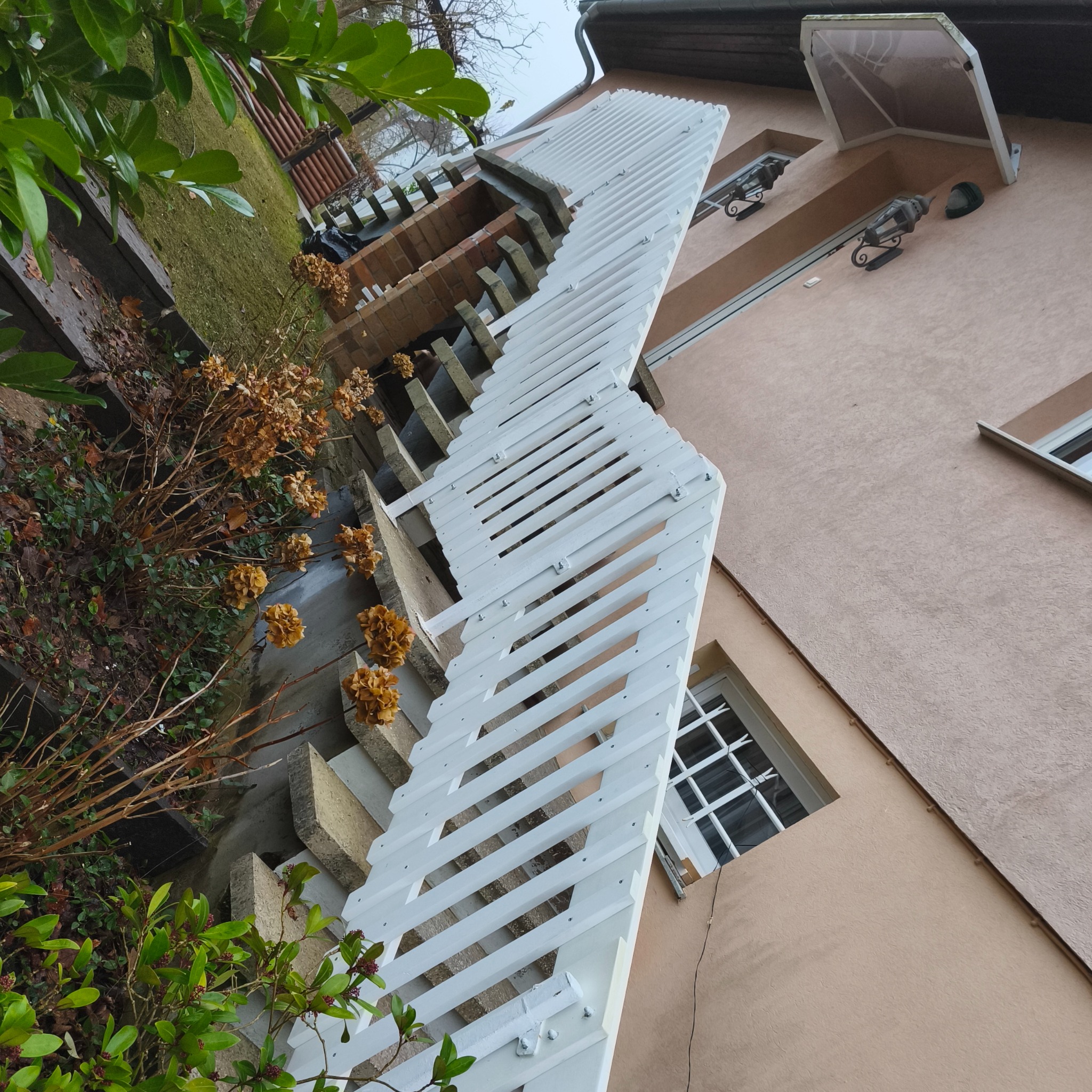 Biała, drewniana pergola nad schodami zewnętrznymi, przymocowana do ściany budynku. Widoczne rośliny i elementy architektury. Ujęcie z dołu, podkreślające konstrukcję.