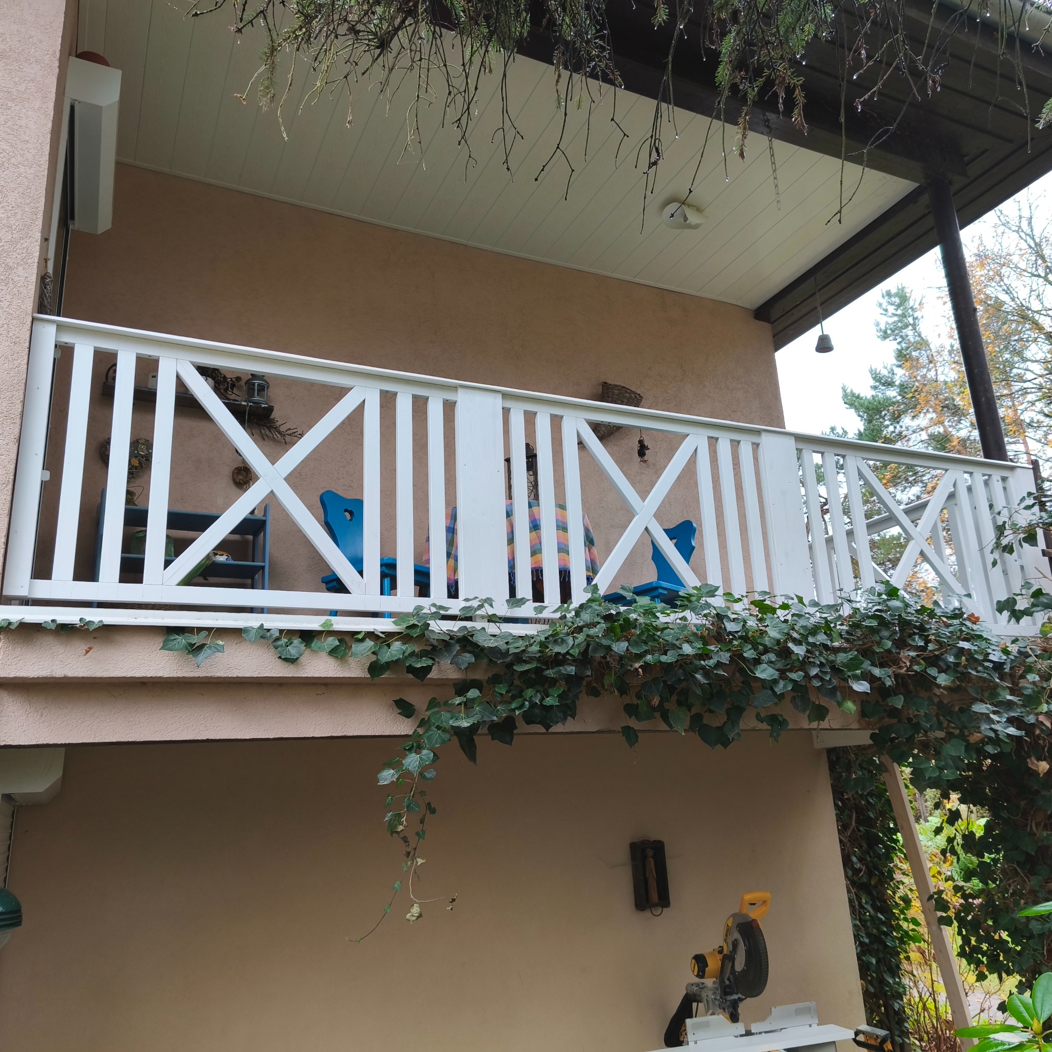 Balkon z białą, drewnianą balustradą w kształcie krzyży, ozdobiony bluszczem. W tle widoczne niebieskie krzesła i dekoracje. Na dole piła do drewna.