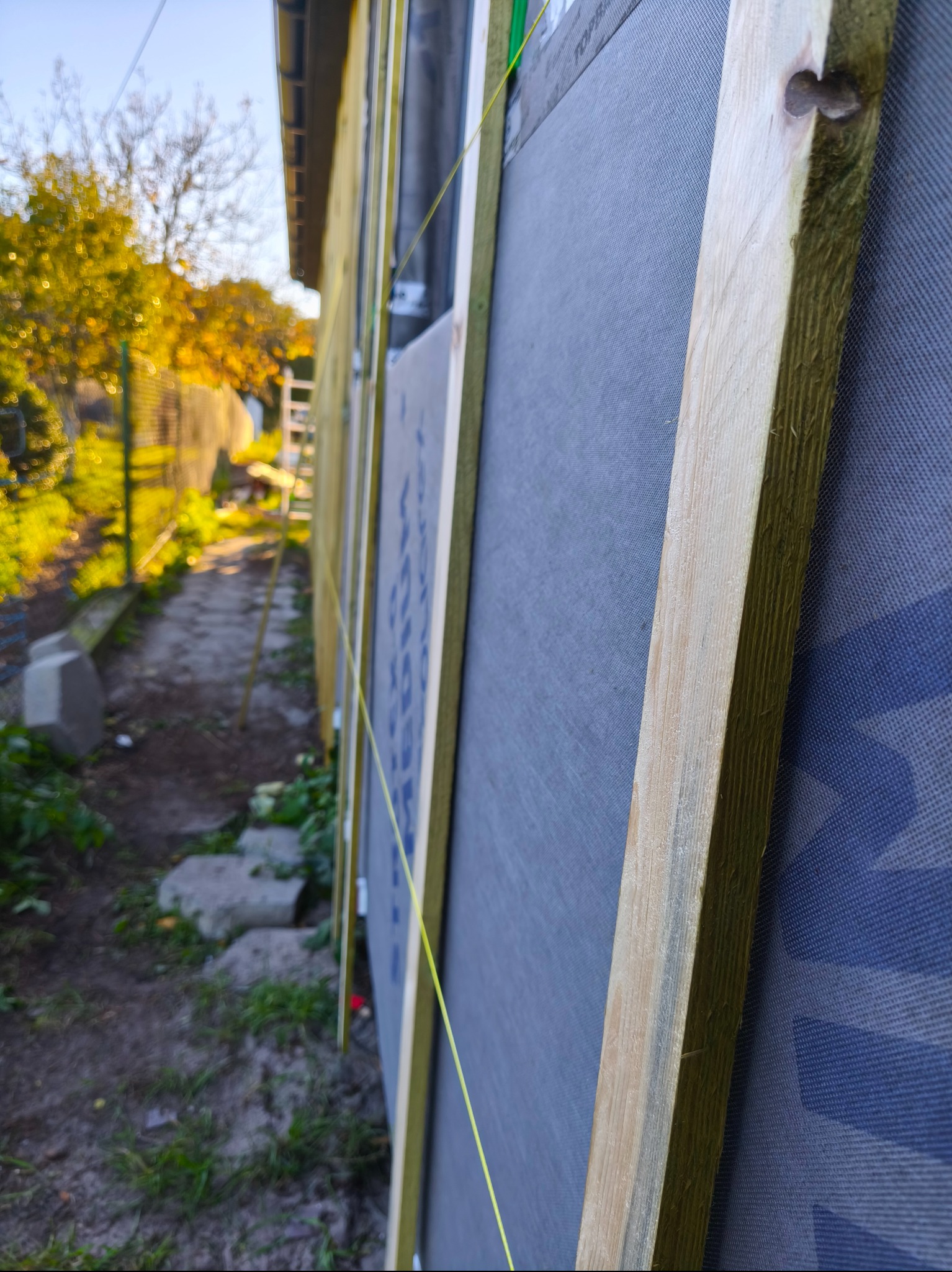 Fragment ściany domu w trakcie budowy, widoczne drewniane elementy konstrukcyjne i szara membrana. W tle zieleń i fragment ogrodzenia. Ujęcie z boku, z bliska.