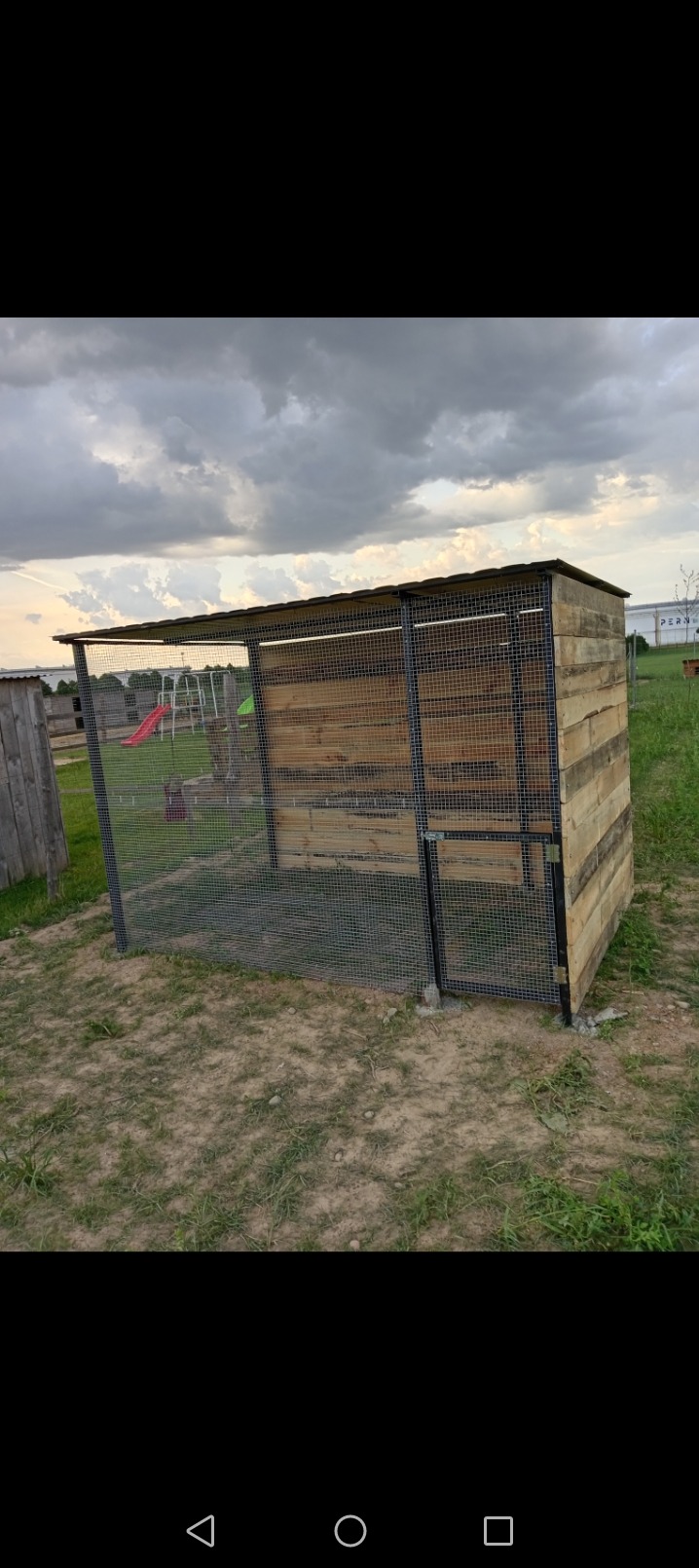 Klatka dla zwierząt z drewnianymi ścianami i metalową siatką, z widocznym placem zabaw w tle, wykonana na zewnątrz w pochmurny dzień.