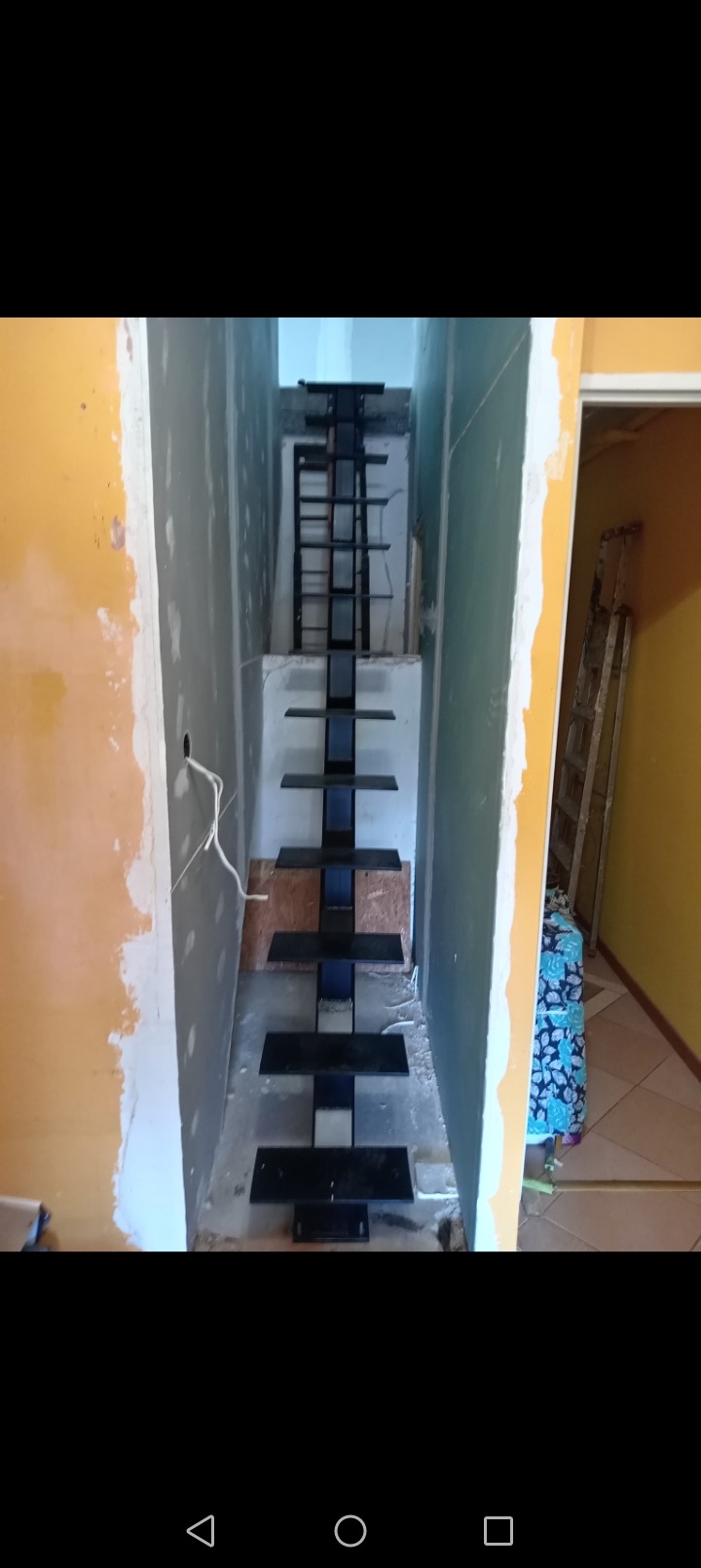 Wąskie schody metalowe bez balustrad, w trakcie montażu w surowym wnętrzu, widoczne niedokończone ściany i przewody elektryczne.