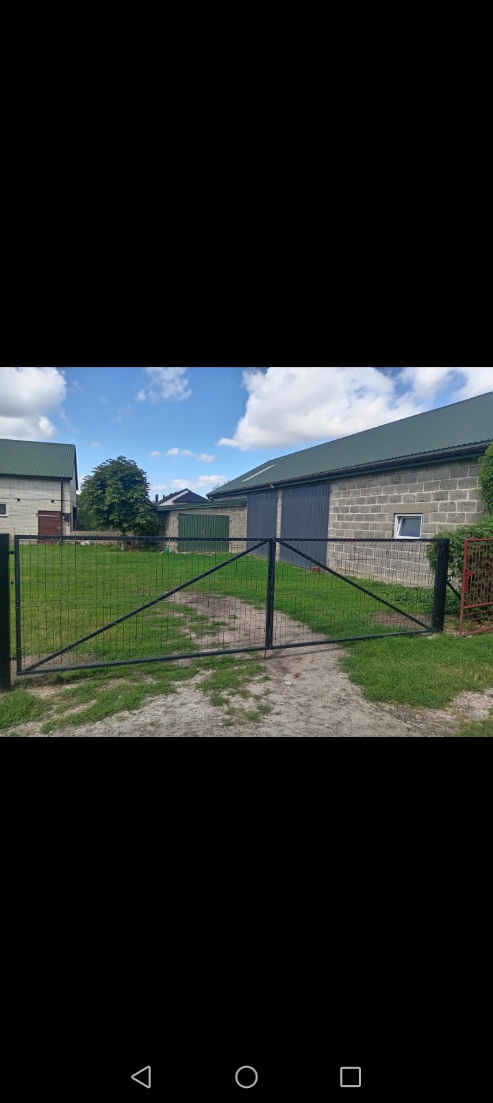 Czarna, metalowa brama wjazdowa z wypełnieniem z siatki na tle budynków gospodarczych i zieleni.