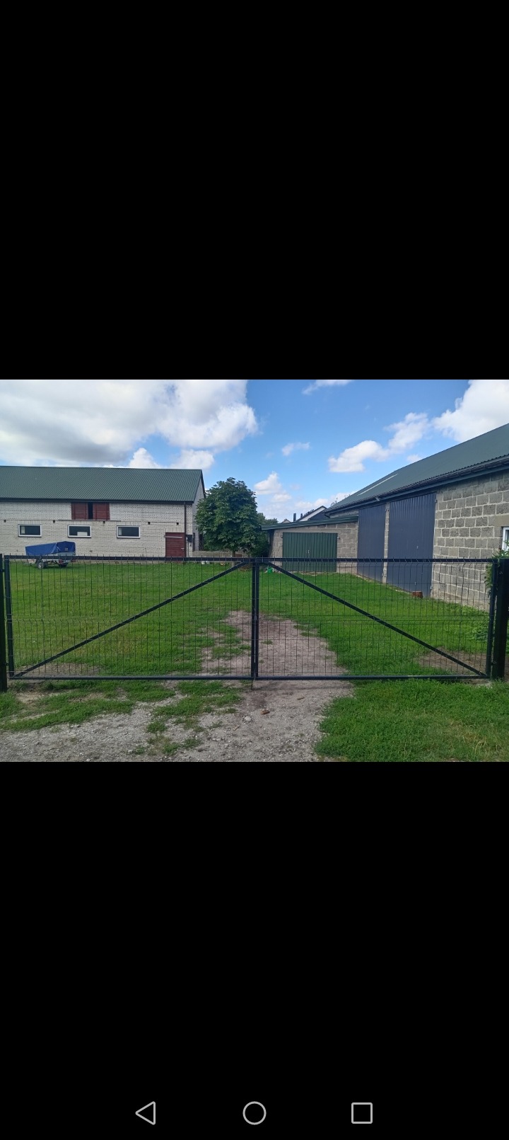 Czarna, metalowa brama panelowa z widoczną konstrukcją wzmacniającą, prowadząca na posesję z budynkami gospodarczymi i fragmentem trawnika.