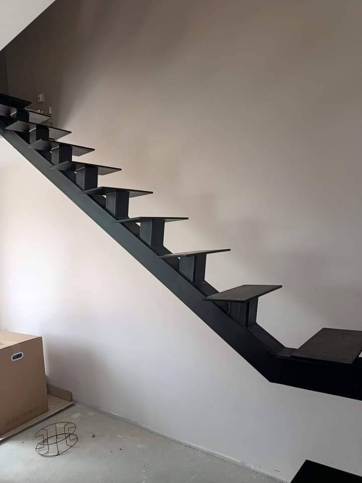 Surowe, metalowe schody bez balustrady, pomalowane na czarno, widoczne na tle białej ściany w trakcie montażu; karton i metalowy element na podłodze wskazują na trwające prace wykończeniowe.