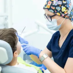 Lekarz w czepku z motywem kwiatowym i okularach, w trakcie badania pacjenta na fotelu dentystycznym, używając metalowego narzędzia. Widoczna maska ochronna i niebieskie rękawiczki.
