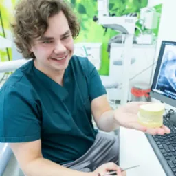 Uśmiechnięty lekarz w zielonym uniformie prezentuje model szczęki na dłoni, w tle monitor z prześwietleniem zębów i sprzęt medyczny.