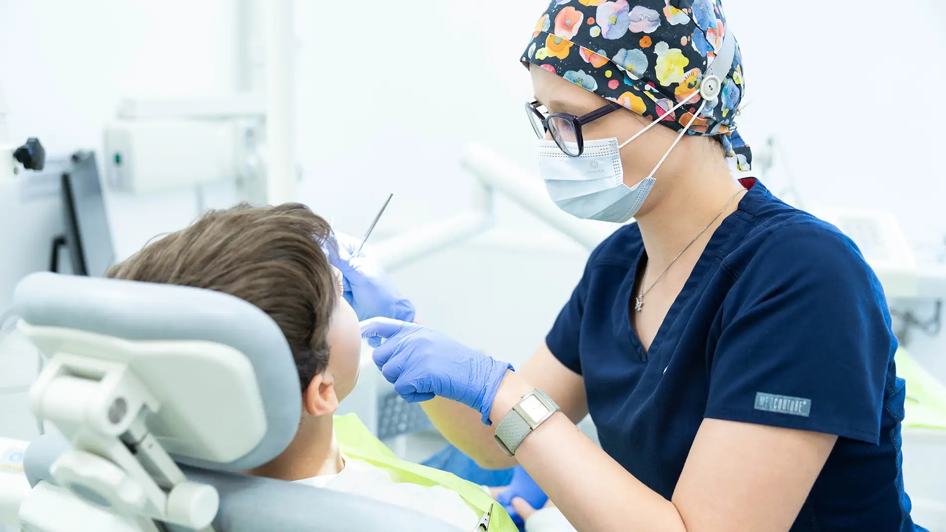 Lekarz w czepku z motywem kwiatowym i okularach, w trakcie badania pacjenta na fotelu dentystycznym, używając metalowego narzędzia. Widoczna maska ochronna i niebieskie rękawiczki.