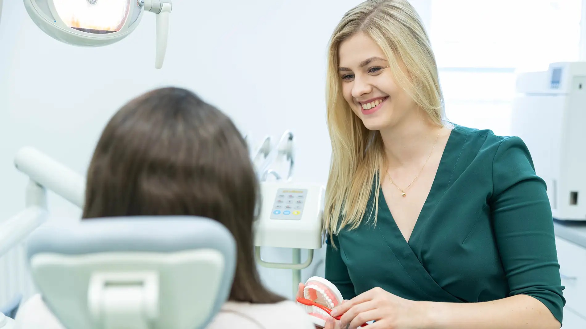 Uśmiechnięta blondynka w zielonym uniformie stomatologicznym demonstruje pacjentowi model szczęki z czerwoną szczoteczką w jasnym gabinecie.