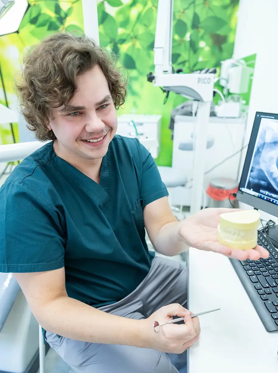 Uśmiechnięty lekarz w zielonym uniformie prezentuje model szczęki na dłoni, w tle monitor z prześwietleniem zębów i sprzęt medyczny.