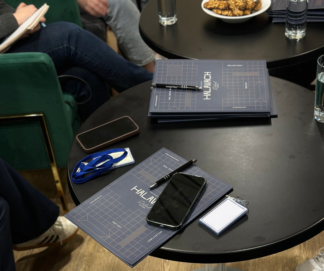 Stolik z materiałami informacyjnymi agencji nieruchomości Halavach Real Estate, telefonami i identyfikatorami. Widoczne osoby w tle, sugerujące spotkanie.