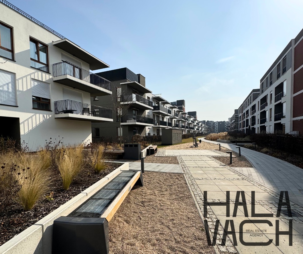 Nowoczesne osiedle z apartamentami, widok z poziomu ulicy z elementami małej architektury. W tle budynki z balkonami i zielenią. Słoneczny dzień.