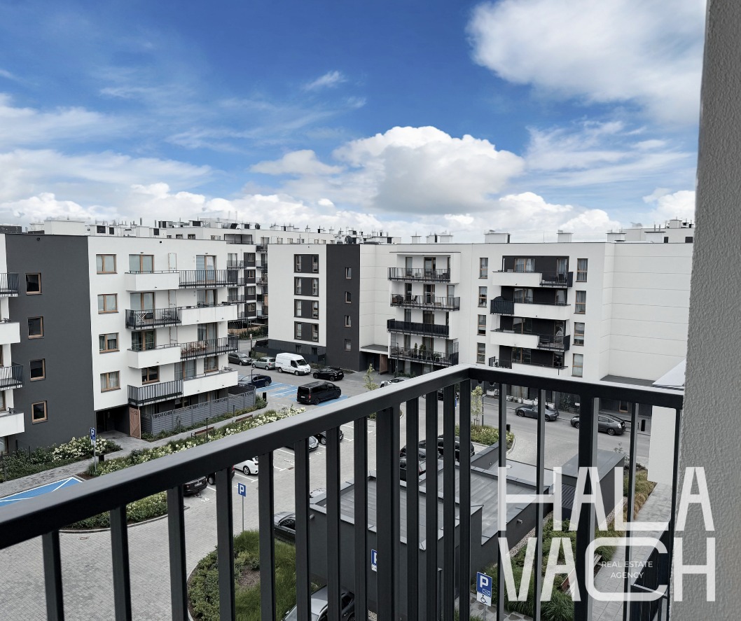 Nowoczesne osiedle z balkonem i widokiem na parking oraz błękitne niebo z chmurami. Ujęcie z wyższego piętra, widoczna barierka balkonu.