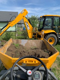Żółta koparko-ładowarka JCB podczas prac ziemnych; widok z kabiny operatora na wywrotkę wypełnioną ziemią, w tle dom i zieleń.