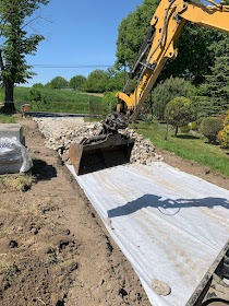 Żółta koparka nabiera gruz na utwardzonej geowłókniną drodze gruntowej, w tle zieleń drzew i błękitne niebo.