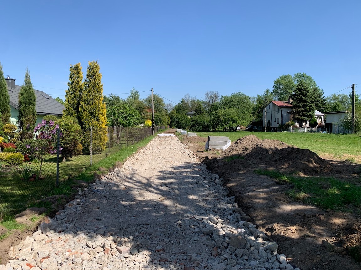 Przygotowanie podłoża drogi gruntowej z użyciem gruzu i kamienia, widoczne wykopane krawędzie i fragmenty domów w tle.