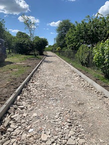 Wąska droga gruntowa w trakcie przygotowania, z ułożonymi obrzeżami betonowymi po bokach, wypełniona gruzem i kamieniami, otoczona zielenią drzew i krzewów w słoneczny dzień.